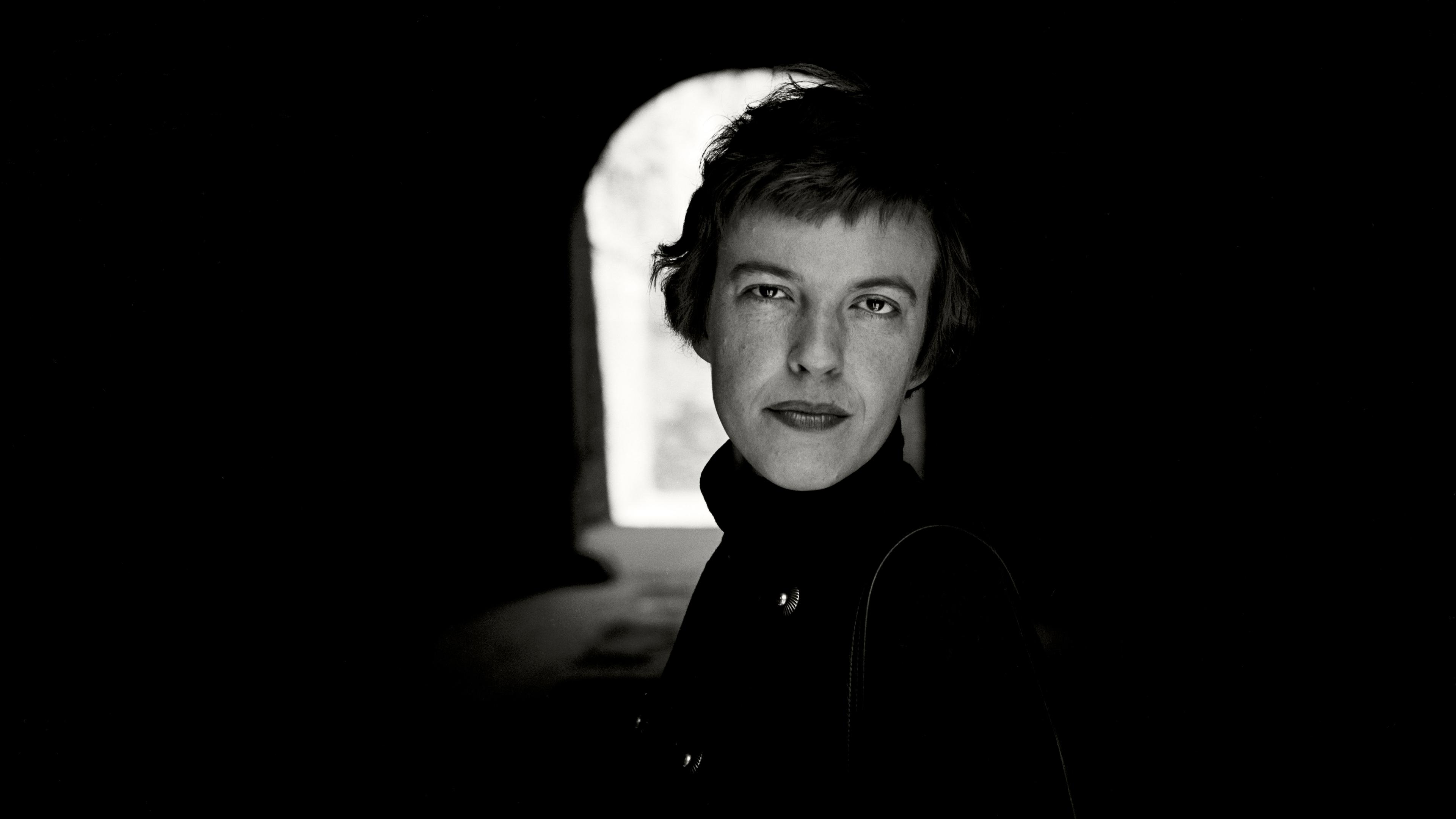 Black and white photo of a person in a dark tunnel with light in the background, wearing a coat, looking at the camera.