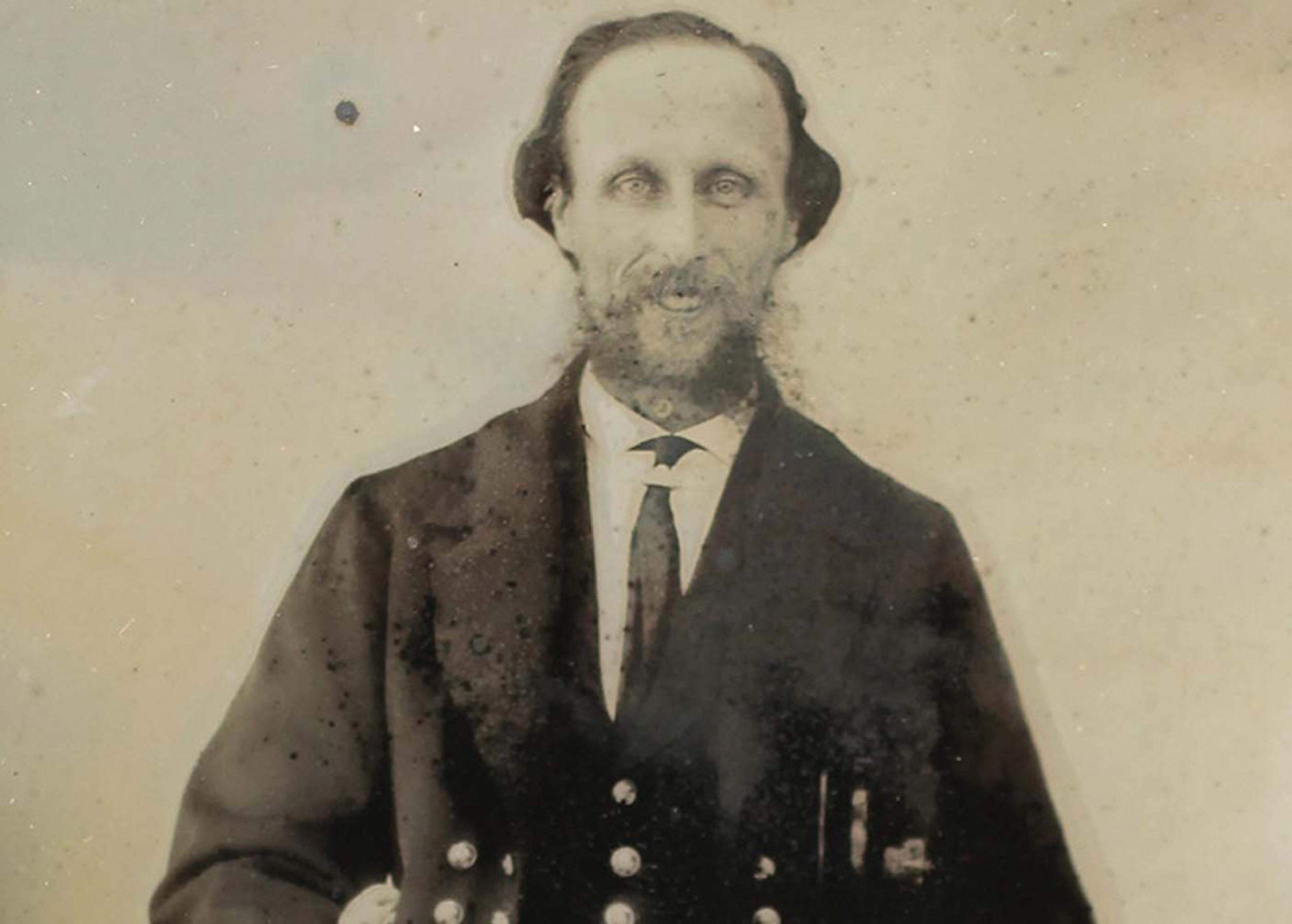 Antique photo of a bearded man in a dark suit with buttons, photo slightly blurred with age and blemishes on the image surface.