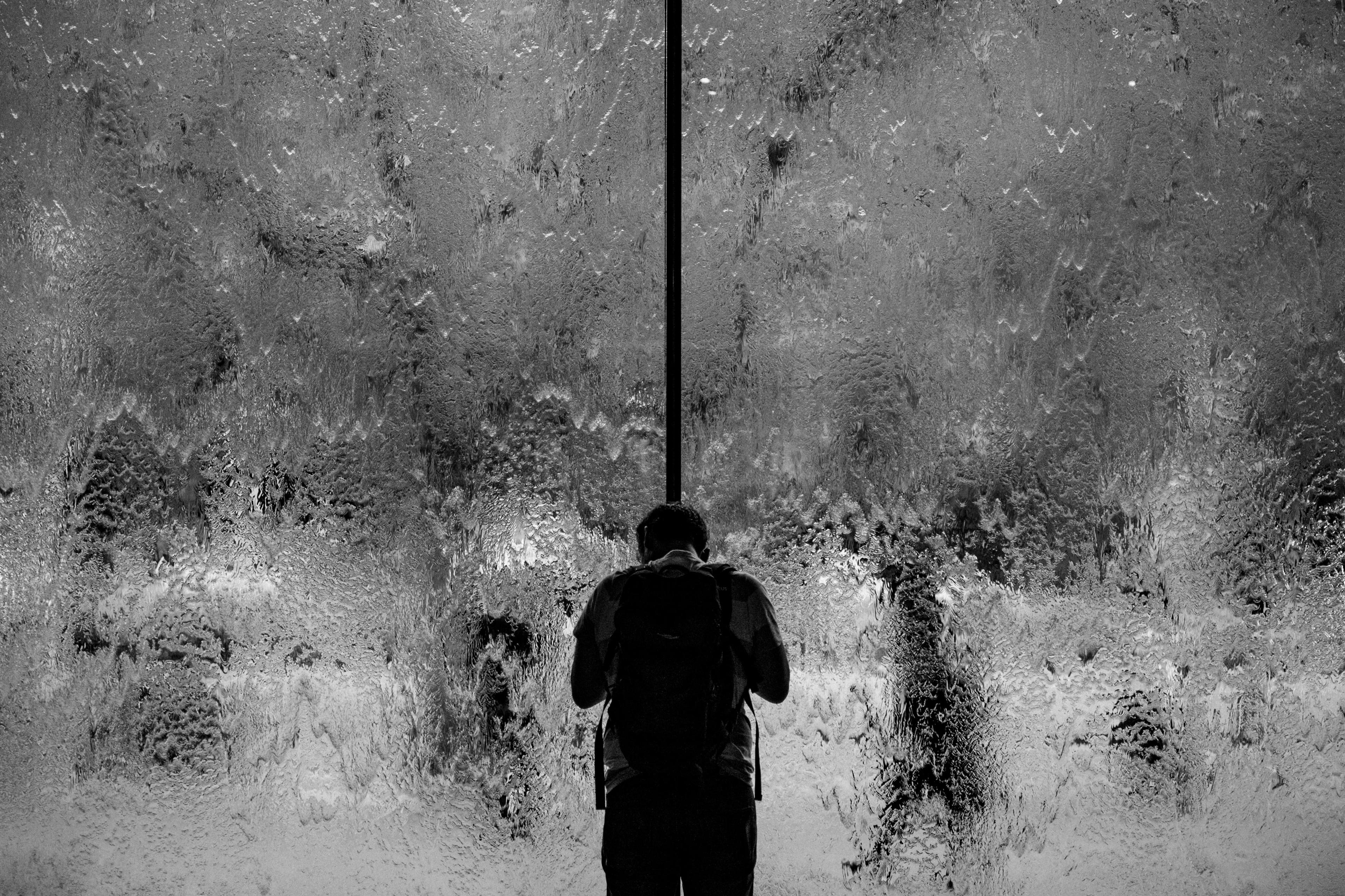 Black and white photo of a person with a backpack standing in front of a textured glass surface, appearing to look through it.