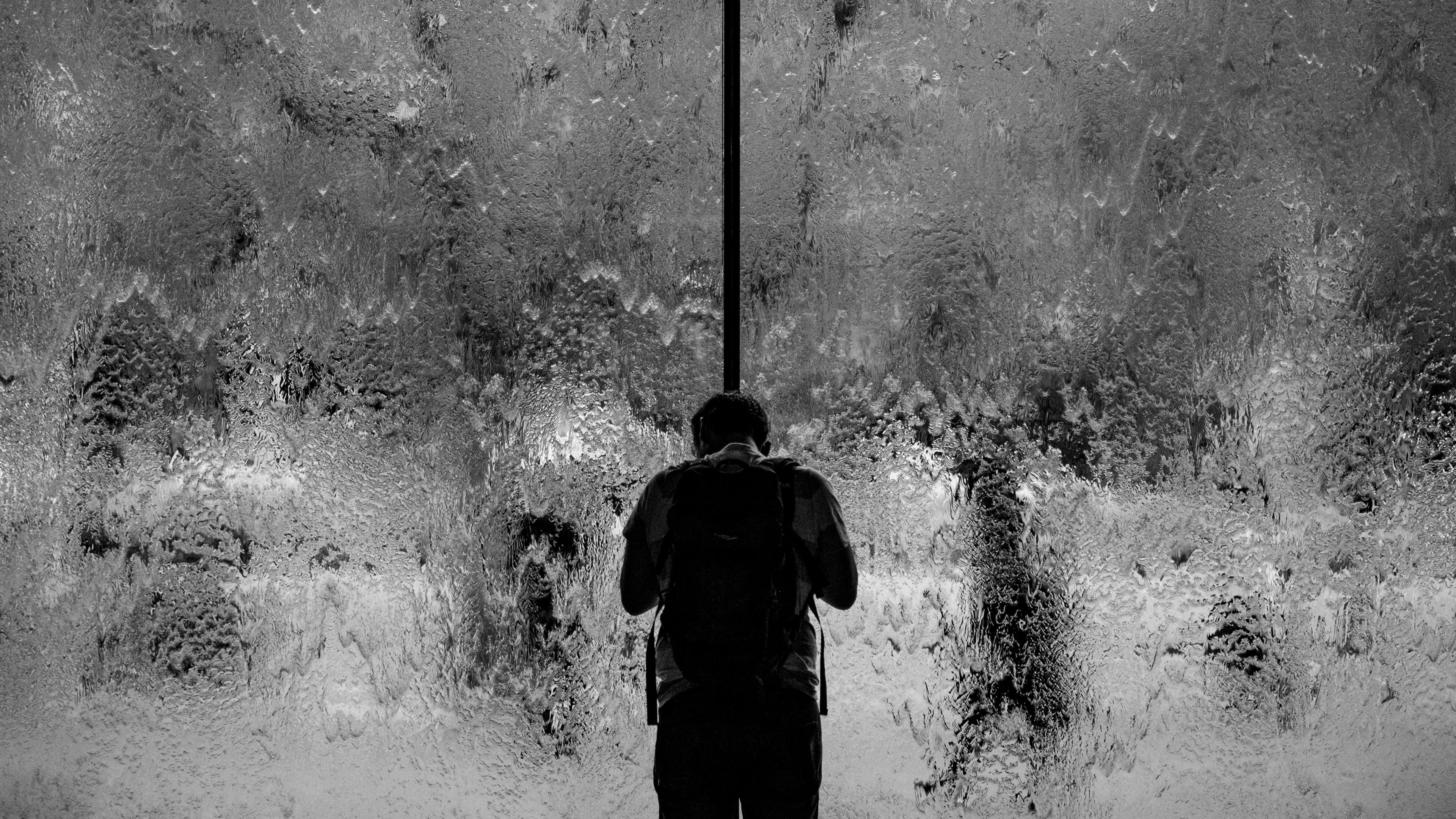 Black and white photo of a person with a backpack standing in front of a textured glass surface, appearing to look through it.