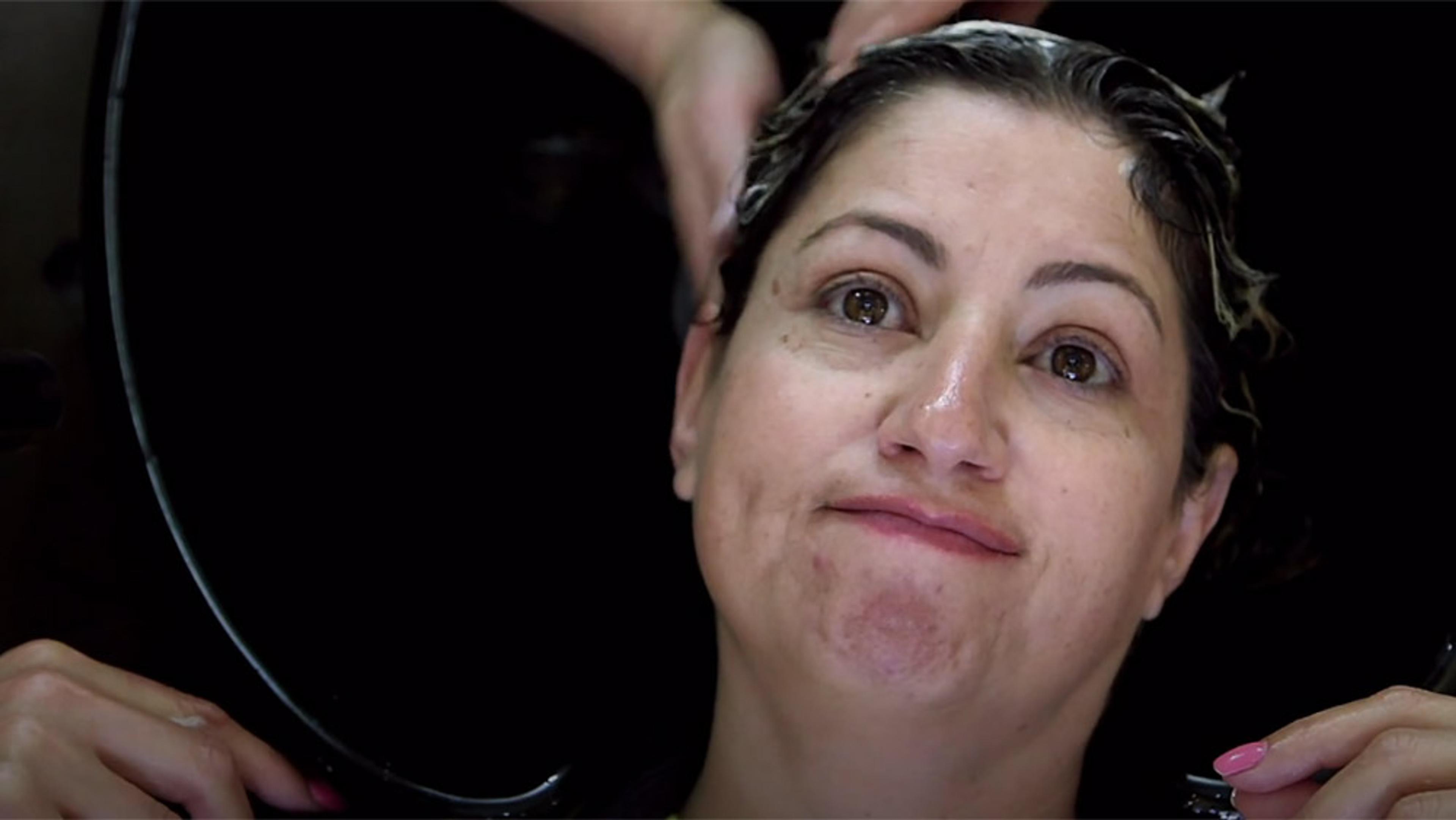 A woman with wet hair sitting in a hairdresser’s basin, getting her hair washed, holding the basin, looking up and smiling slightly.