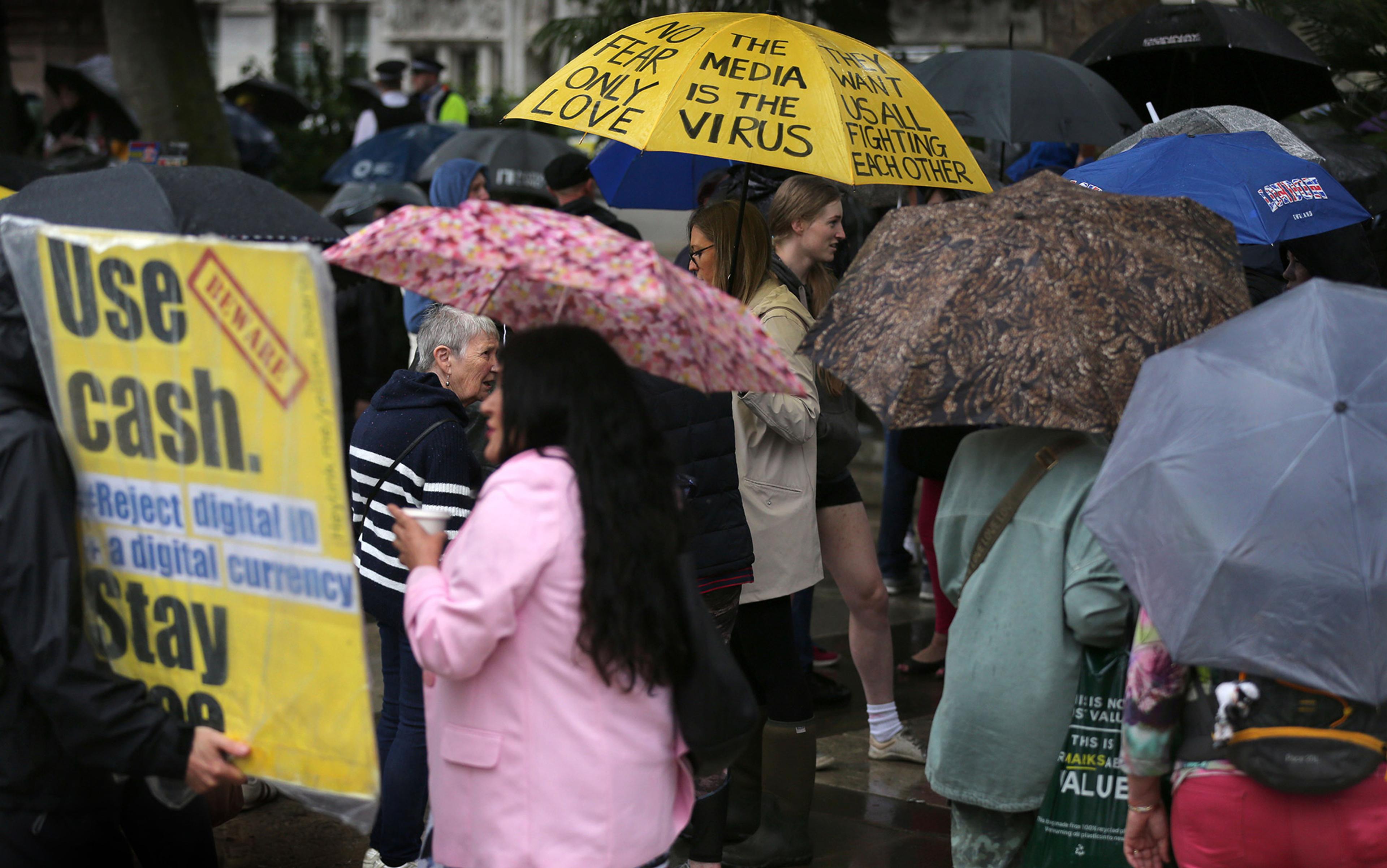A woman in a pink coat and carrying a pink umbrella walks past a protestor carrying a yellow placard appealing to people to use cash