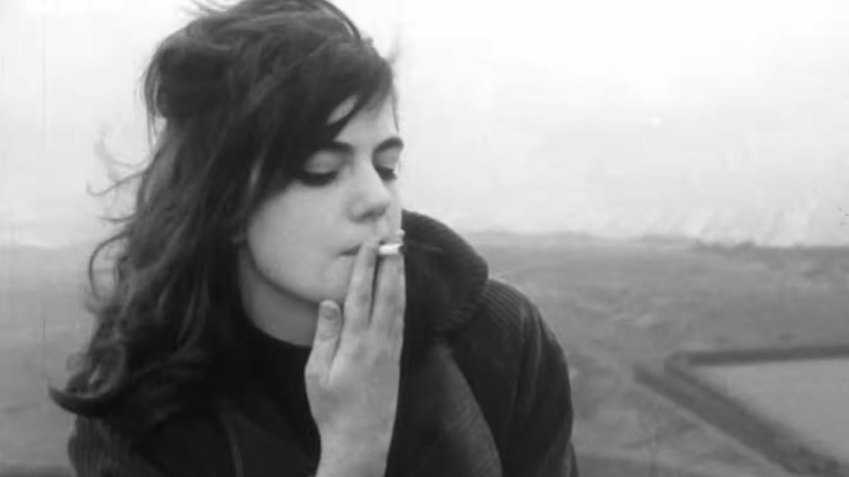 Black and white photo of a teenage girl with dark hair smoking, eyes closed, with a blurred outdoor background.