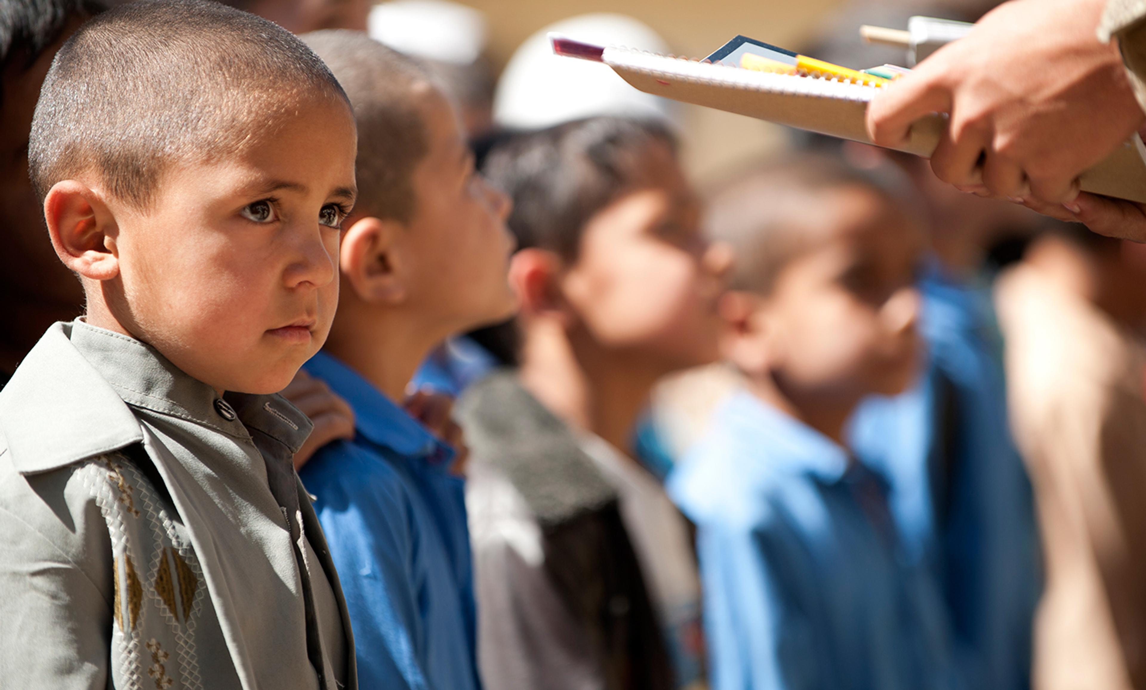 A young boy in line with other children holding school supplies handed out by an adult. The background is out of focus.