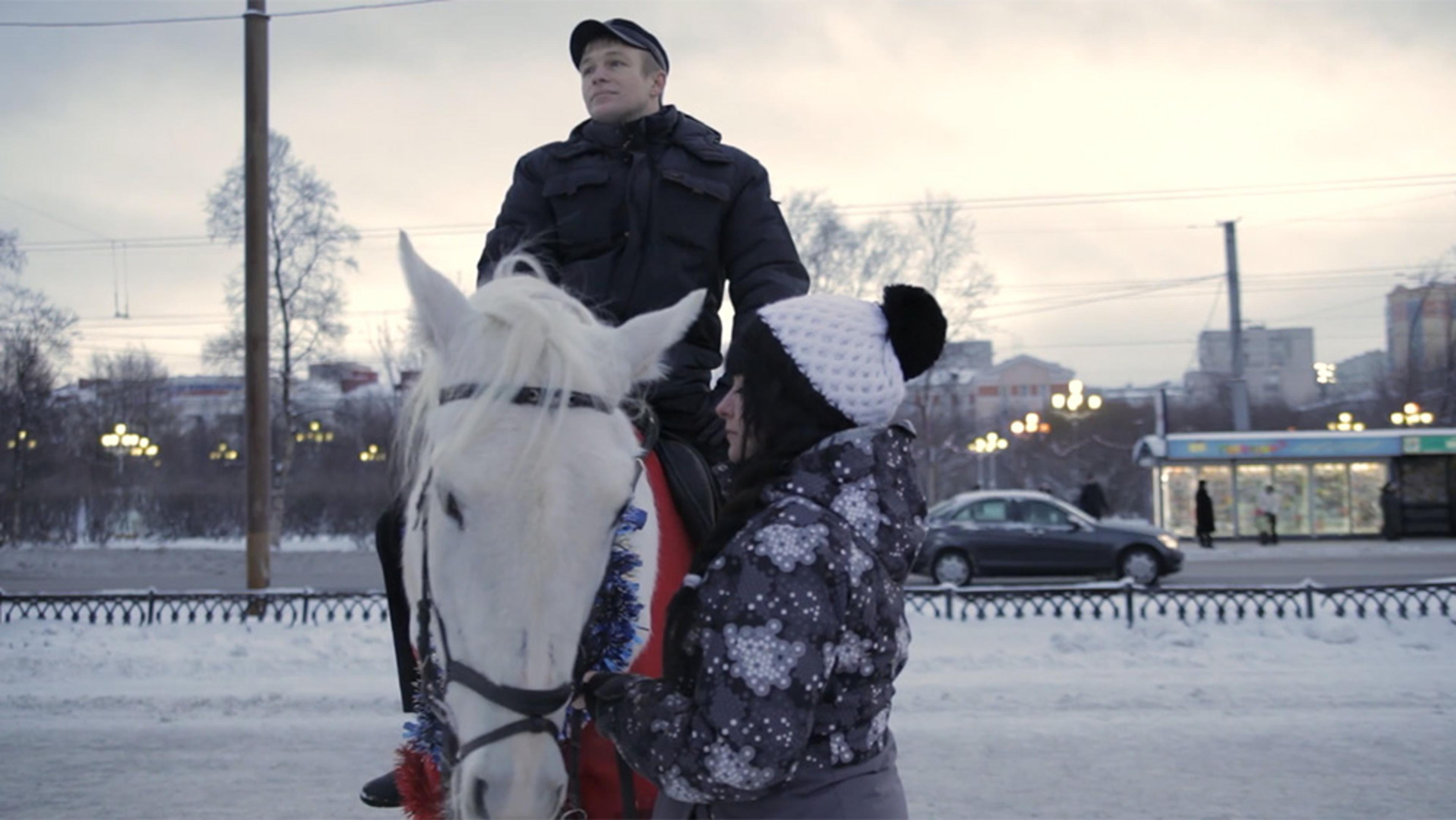 A man on a white horse in a snowy urban setting, with a woman in a winter coat and hat standing by the horse.