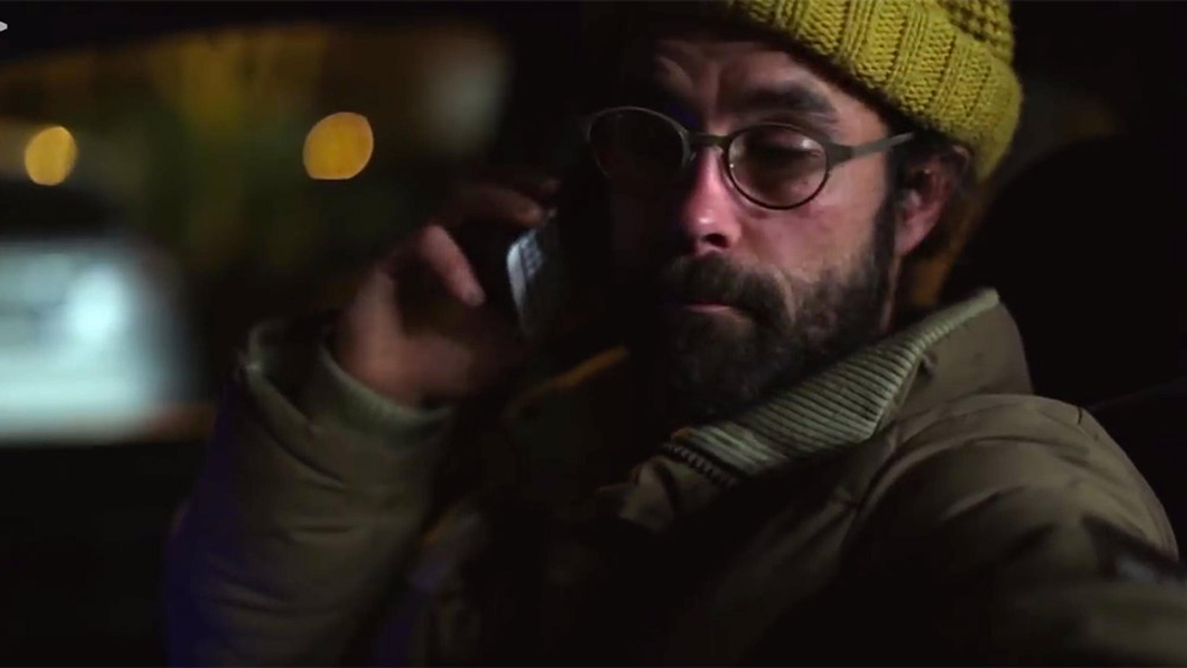 A man with a beard, glasses and a yellow beanie talking on a phone in a dimly lit setting.