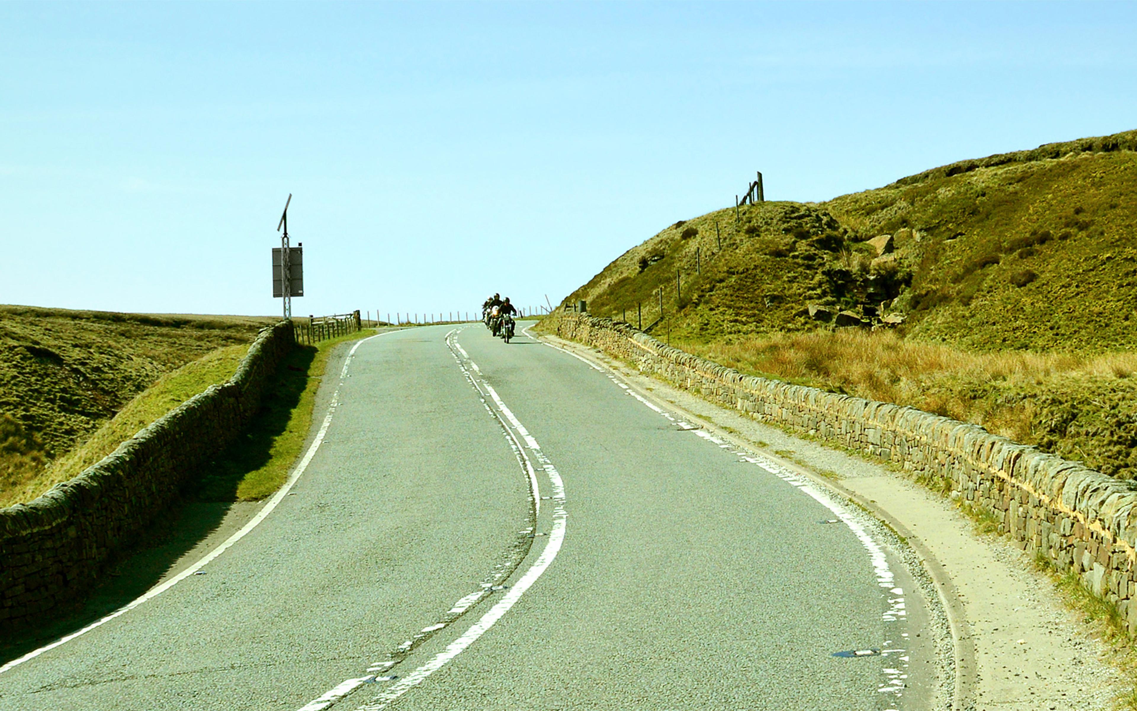 Three motorcyclists are riding on an empty road winding through English moorland beneath a blue sky