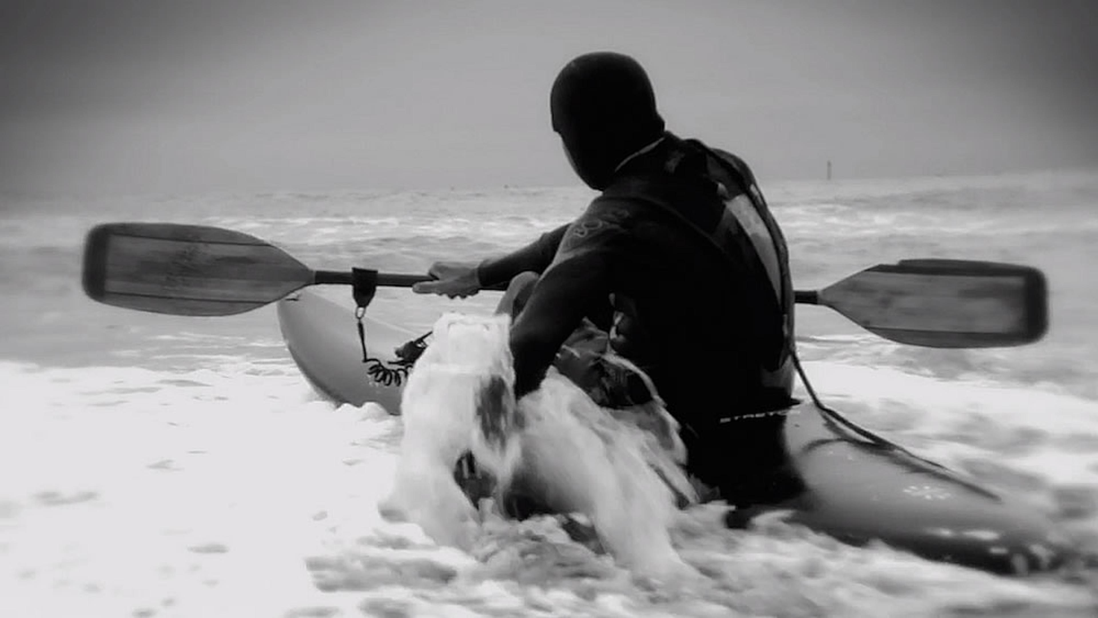 Black and white photo of a man kayaking in the ocean, viewed from behind, paddling through waves on a grey overcast day.