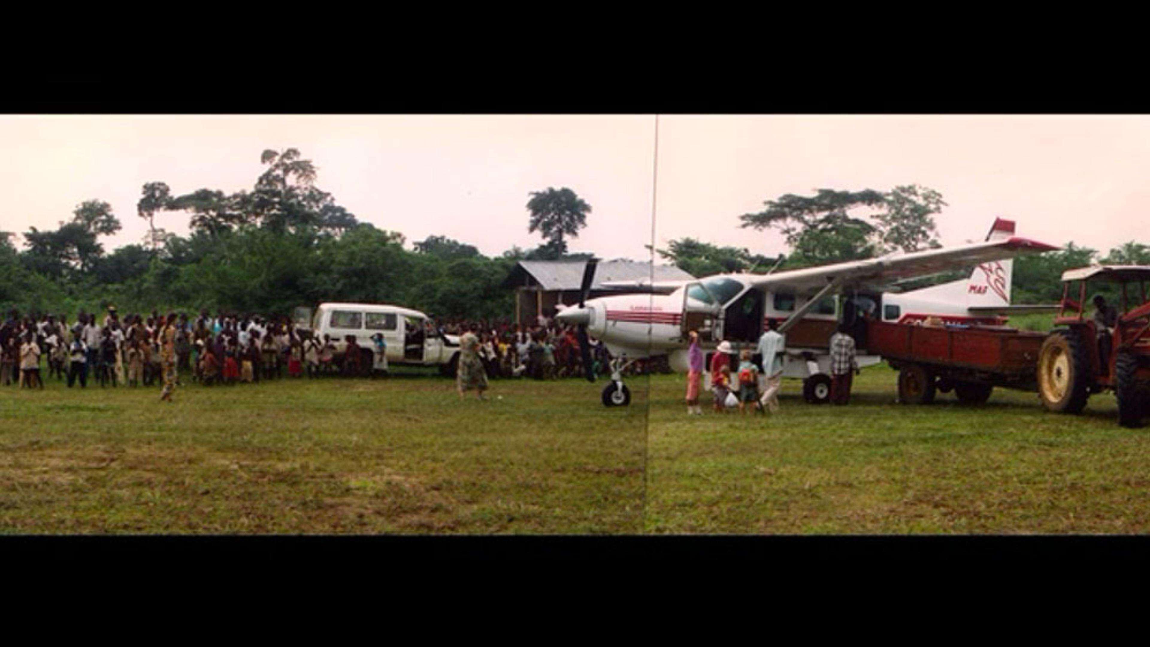 A crowd, a small plane with tribal markings, a van, and a red tractor on a grassy field with trees in the background.