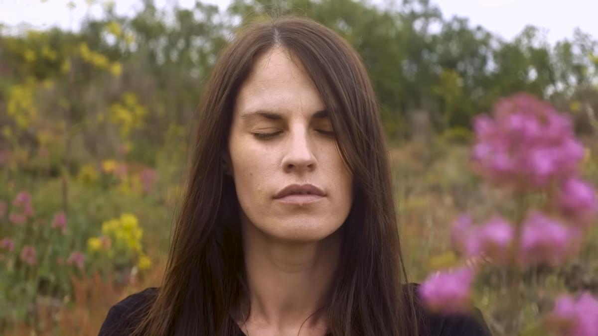 Photo of a woman with long hair, eyes closed in a field of wildflowers and greenery, creating a serene and natural setting.