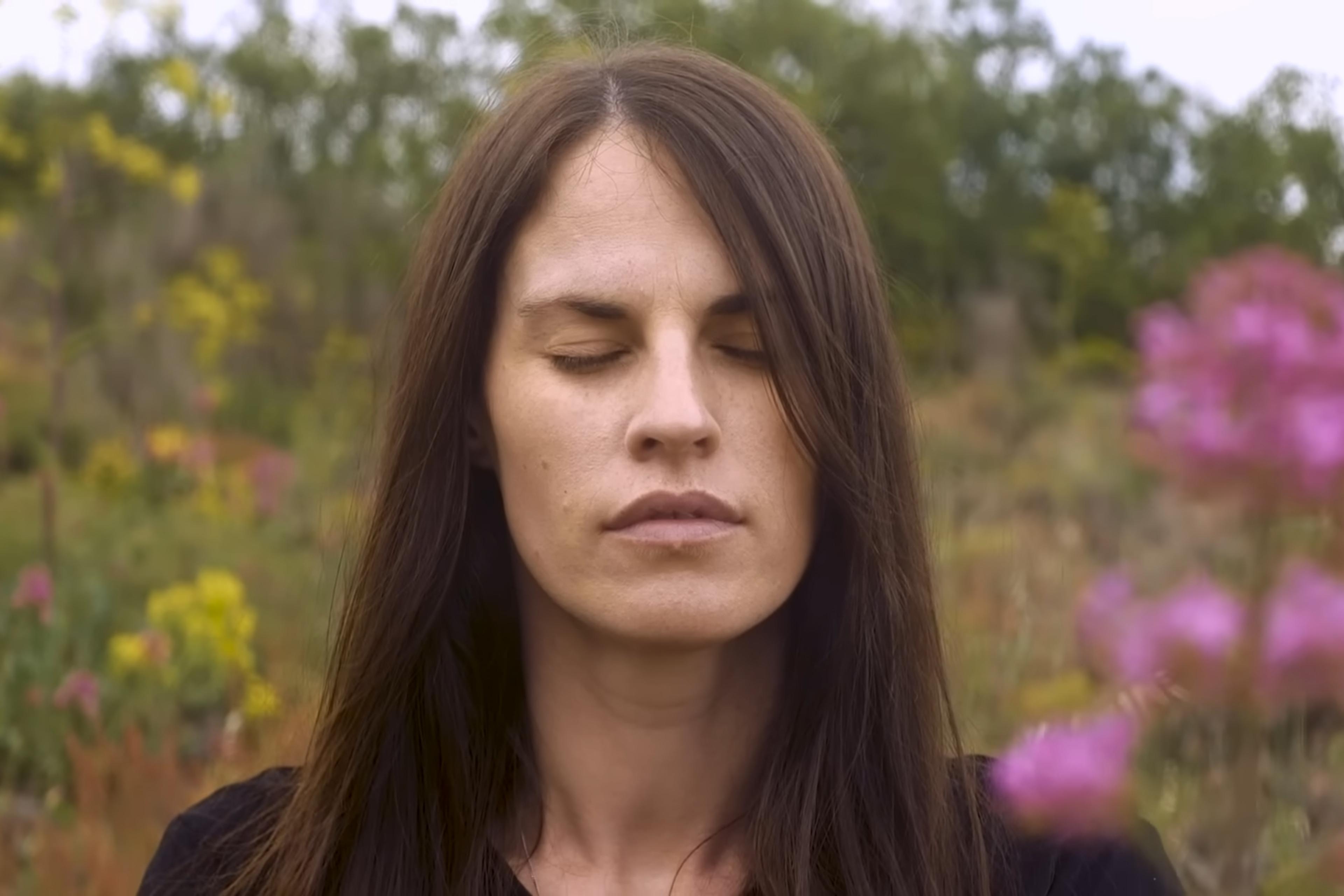 Photo of a woman with long hair, eyes closed in a field of wildflowers and greenery, creating a serene and natural setting.