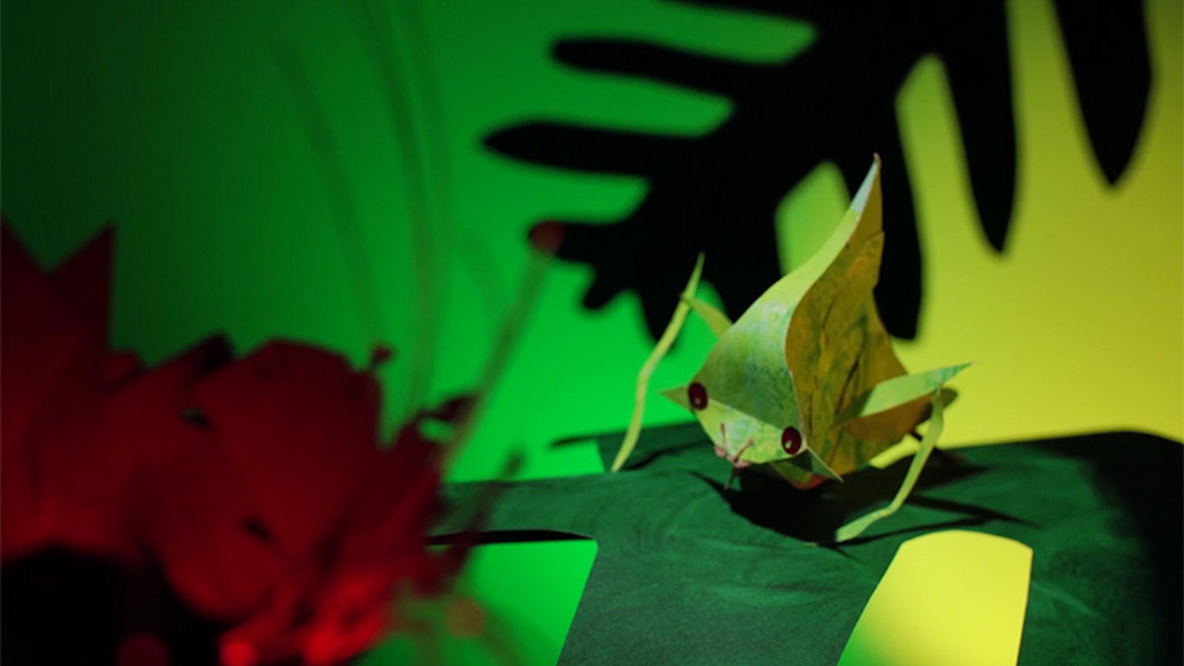 A green origami insect on a dark green leaf with a red flower and a vibrant green-and-yellow background with shadowy leaves.