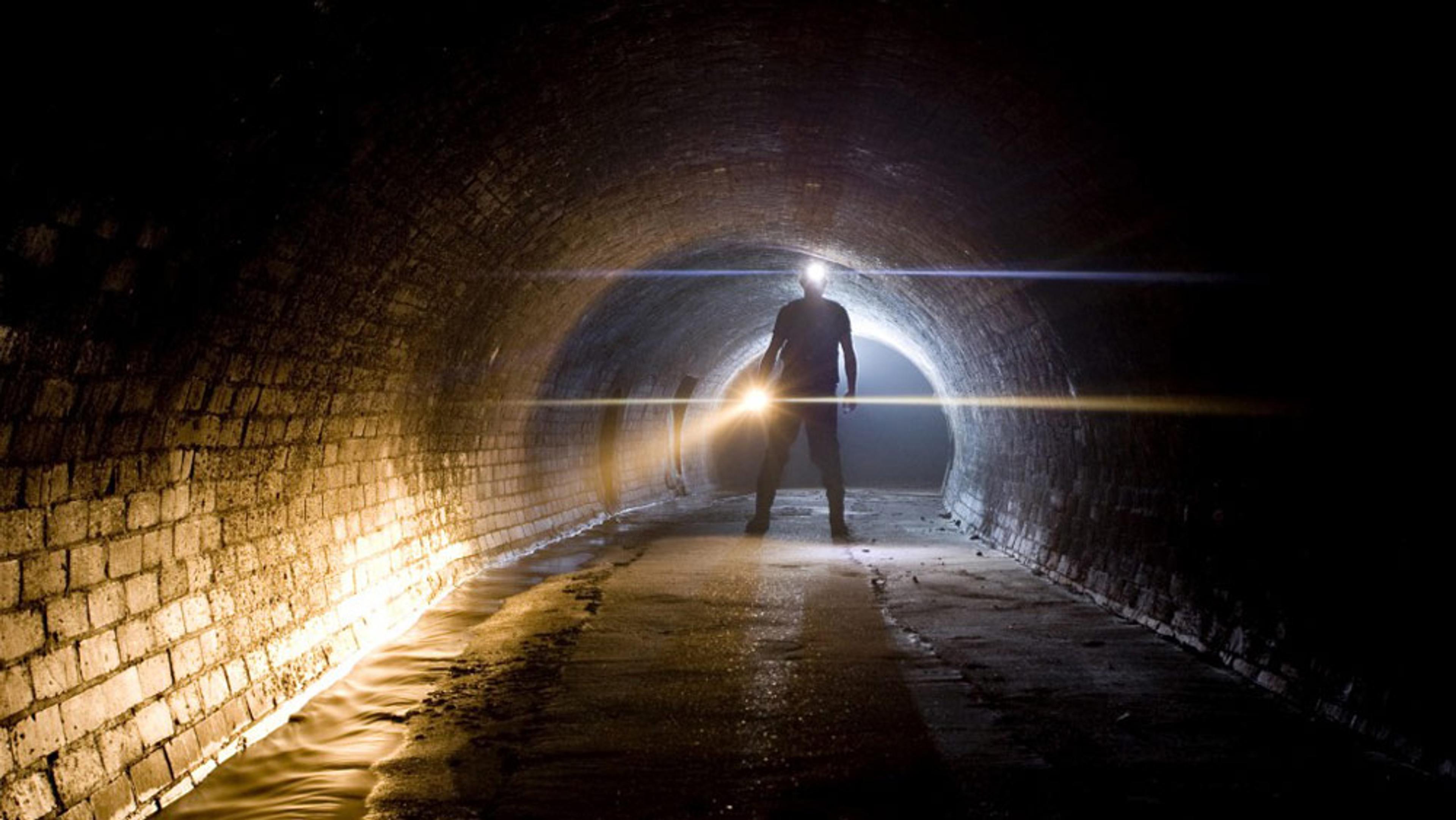 A person standing in a dark tunnel holding a light, with brick walls and a small stream of water running along one side.