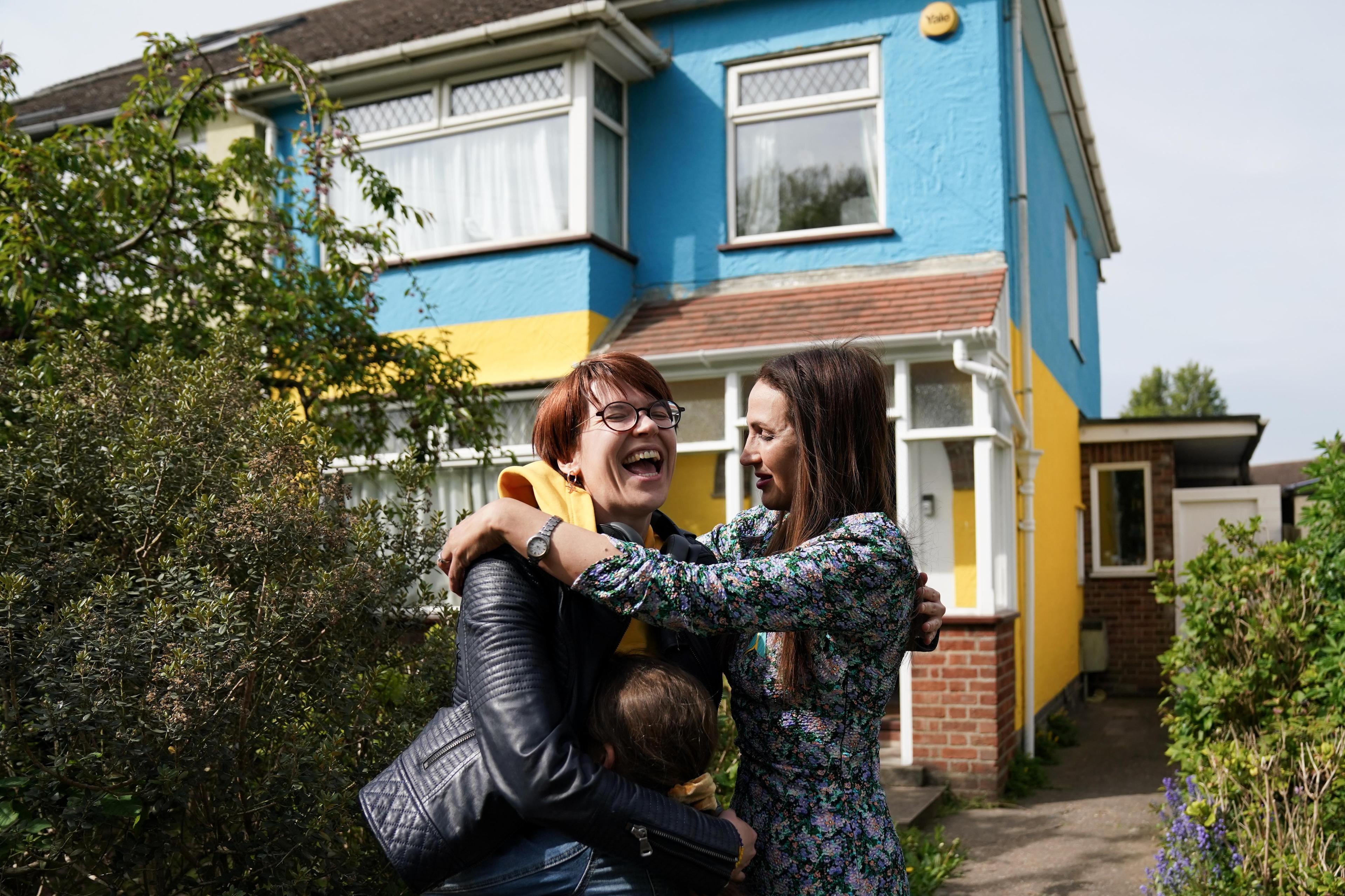 Photo of two women laughing and hugging outside a colourful house with blue and yellow walls, surrounded by greenery.
