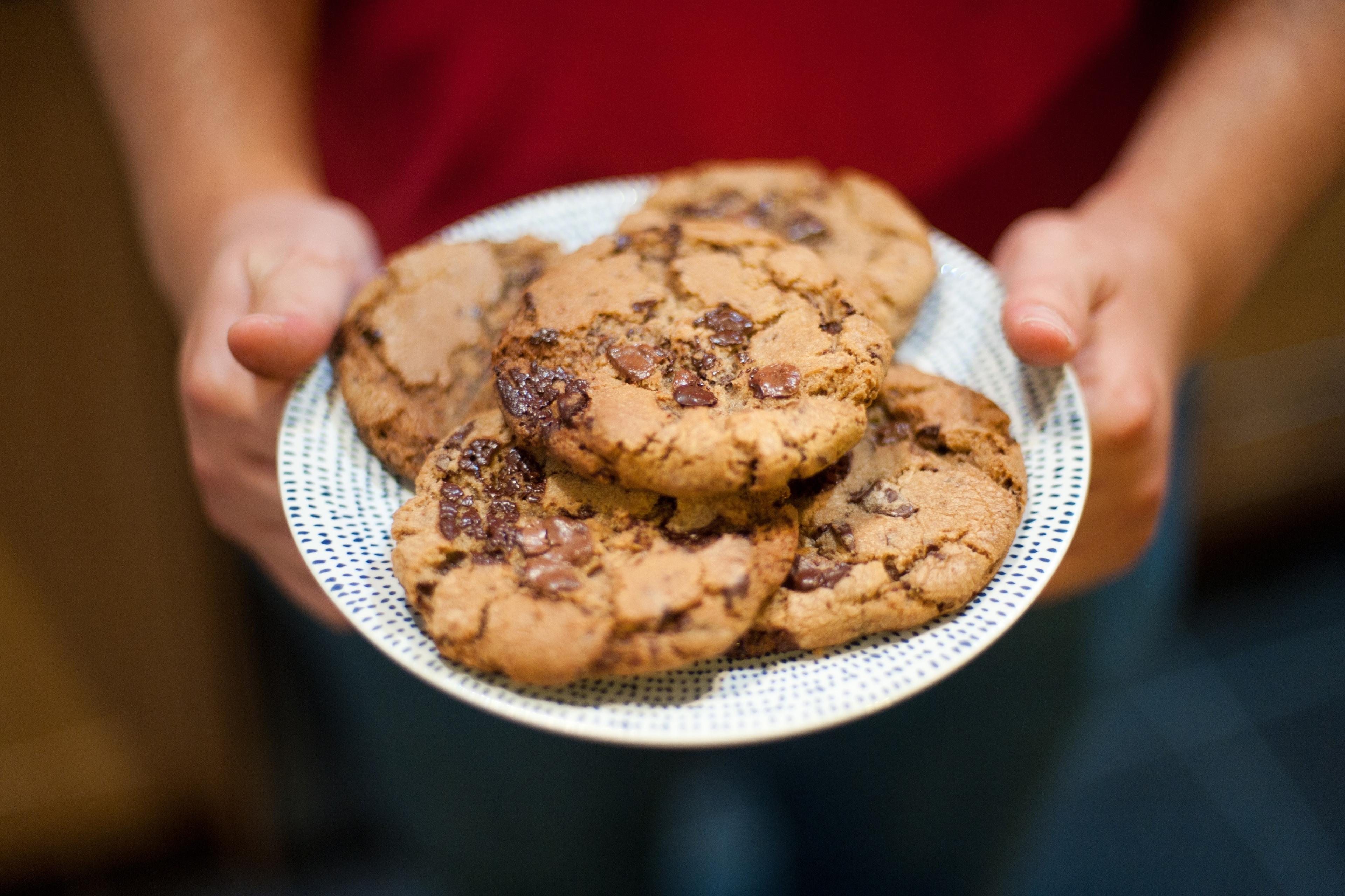 Photo of a person holding a plate with several chocolate chip cookies, set against a blurred background.