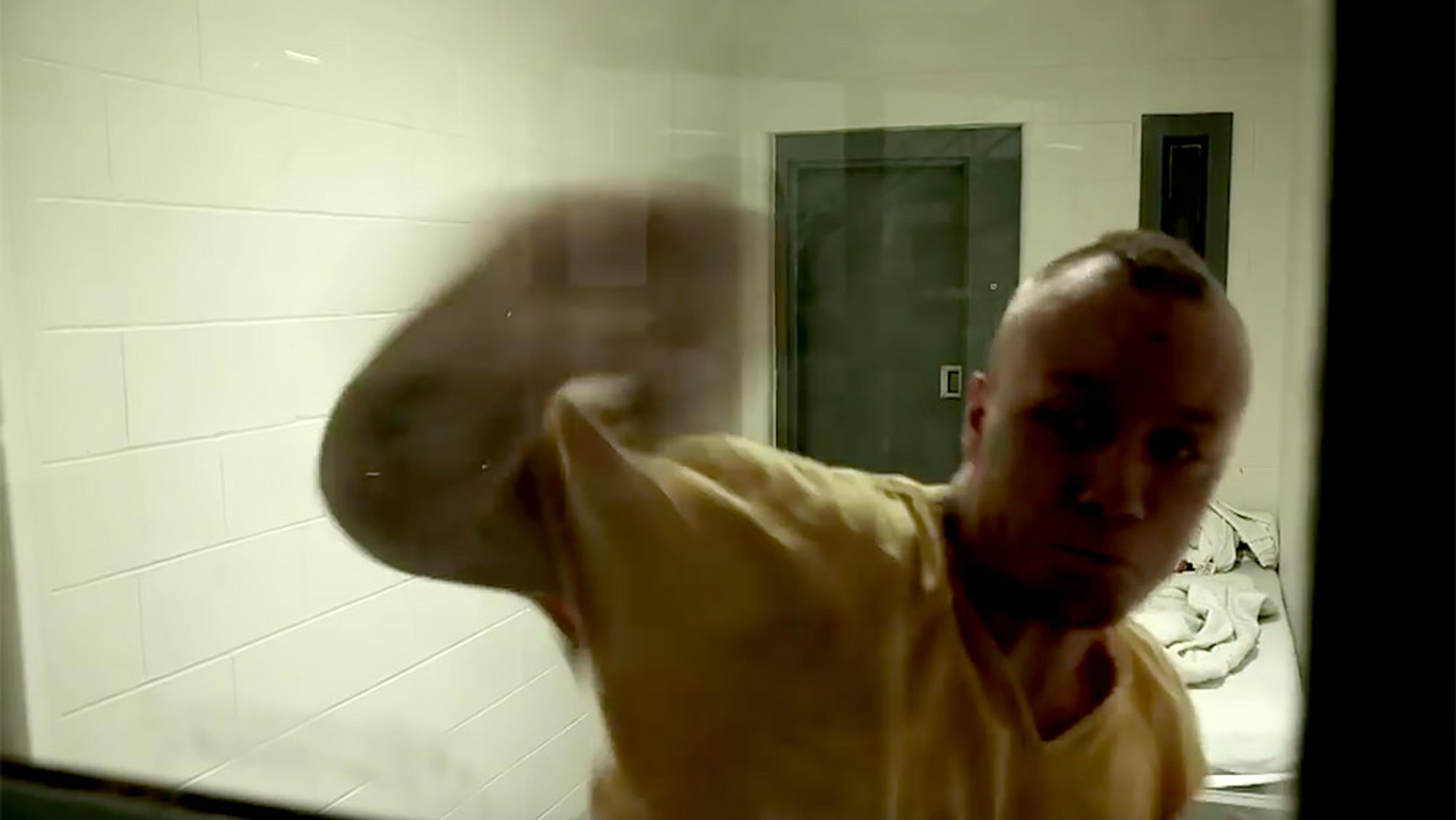 A man in a yellow shirt, in a cell-like room, punching or hitting a glass partition with a bed in the background.
