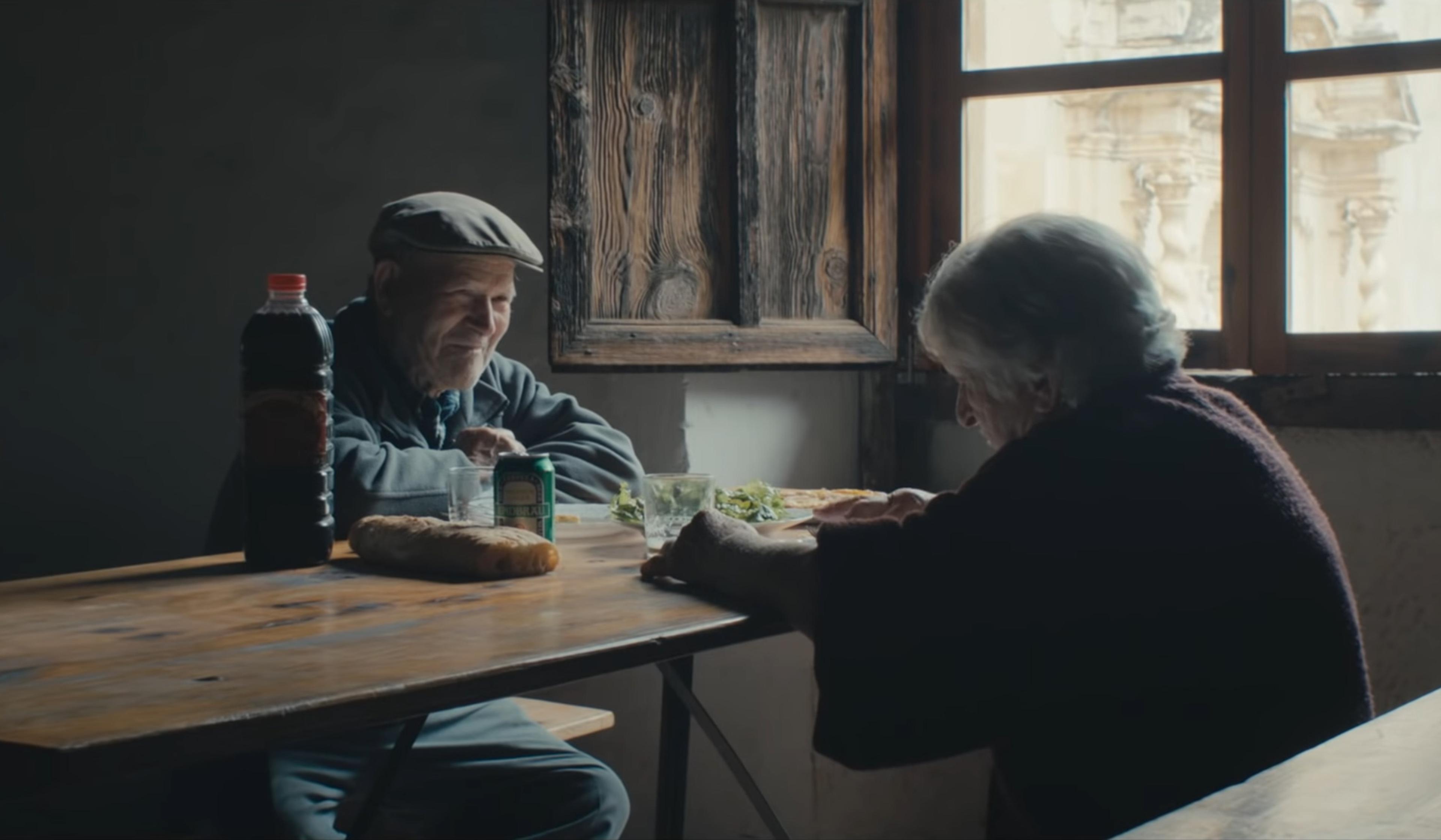 An elderly couple sitting at a wooden table near a window, sharing a meal with bread, salad and drinks on the table.