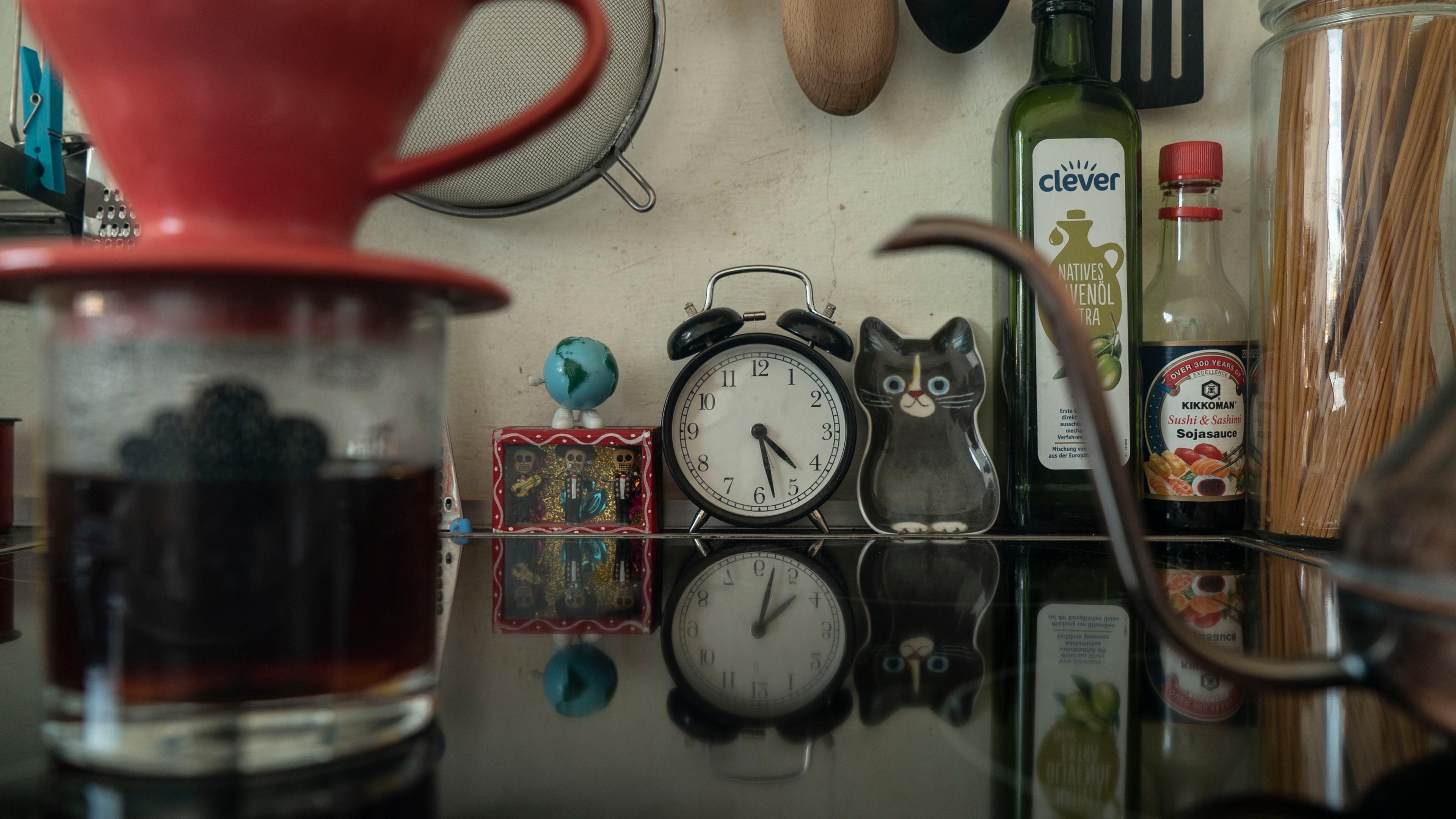 Photo of a kitchen counter with decor, plastic cat, clock and bottles reflected on a glass surface with coffee brewing.