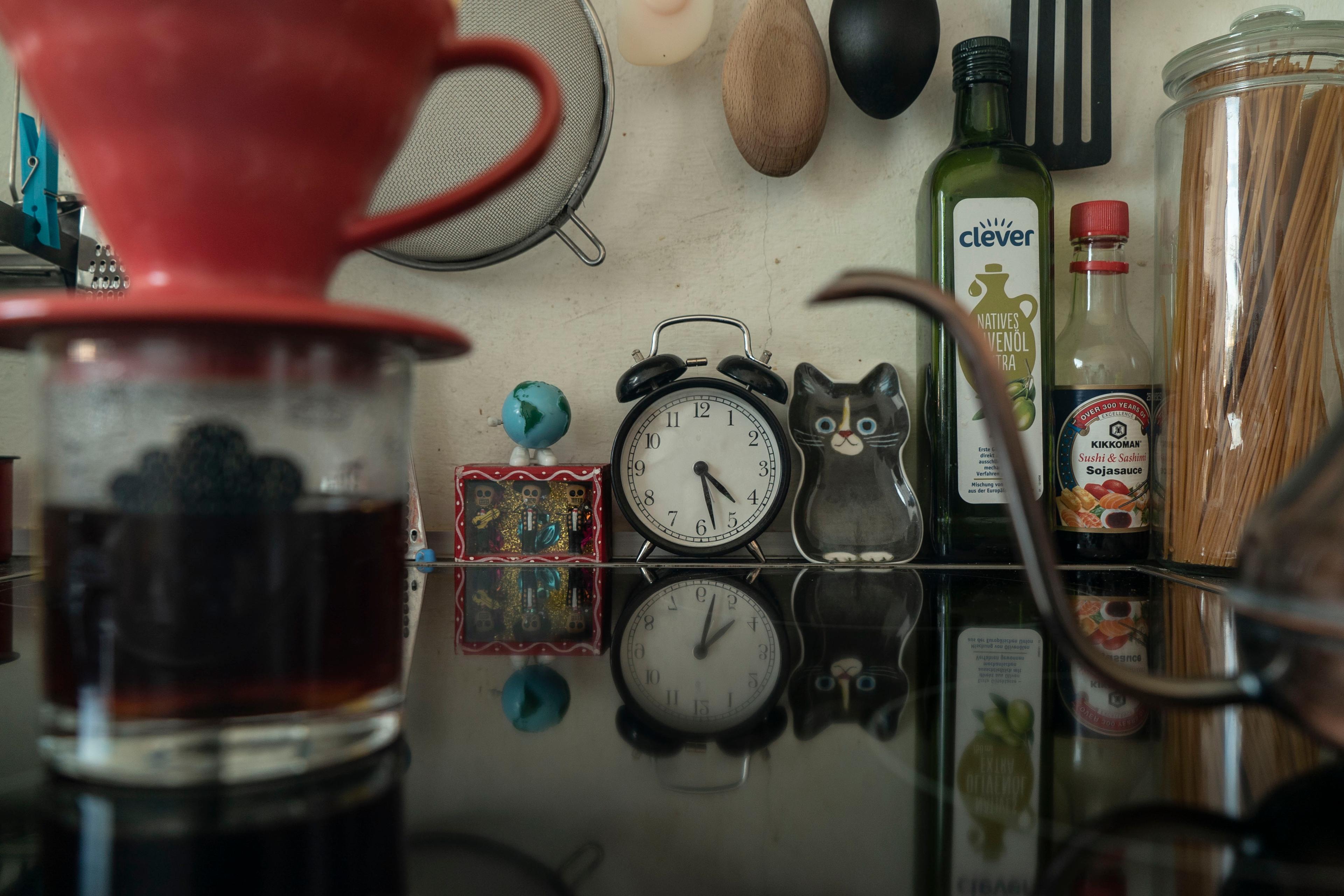 Photo of a kitchen counter with decor, plastic cat, clock and bottles reflected on a glass surface with coffee brewing.