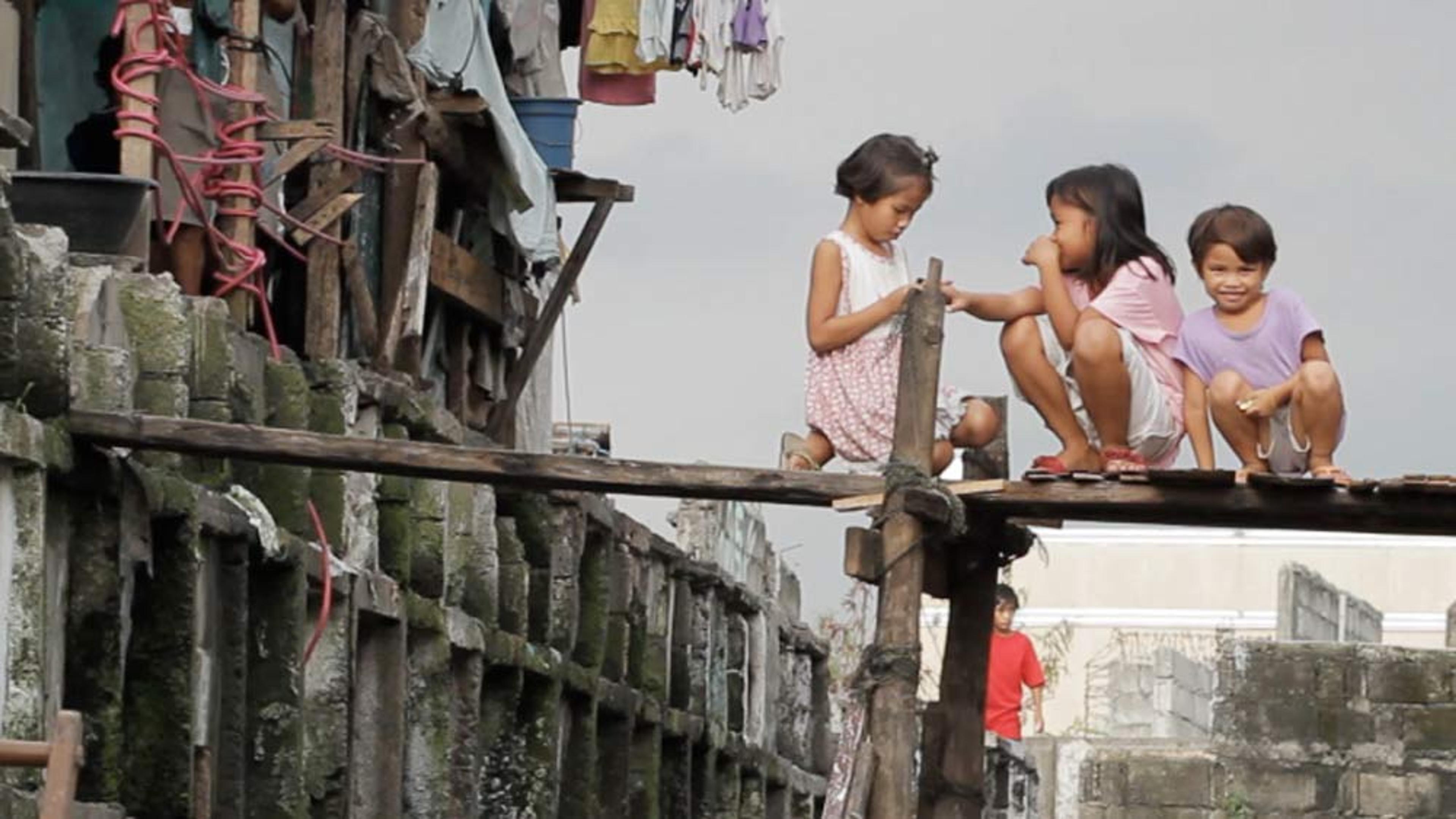 Three children playing on a wooden walkway in a worn-down urban area with buildings and clothes hanging behind them.