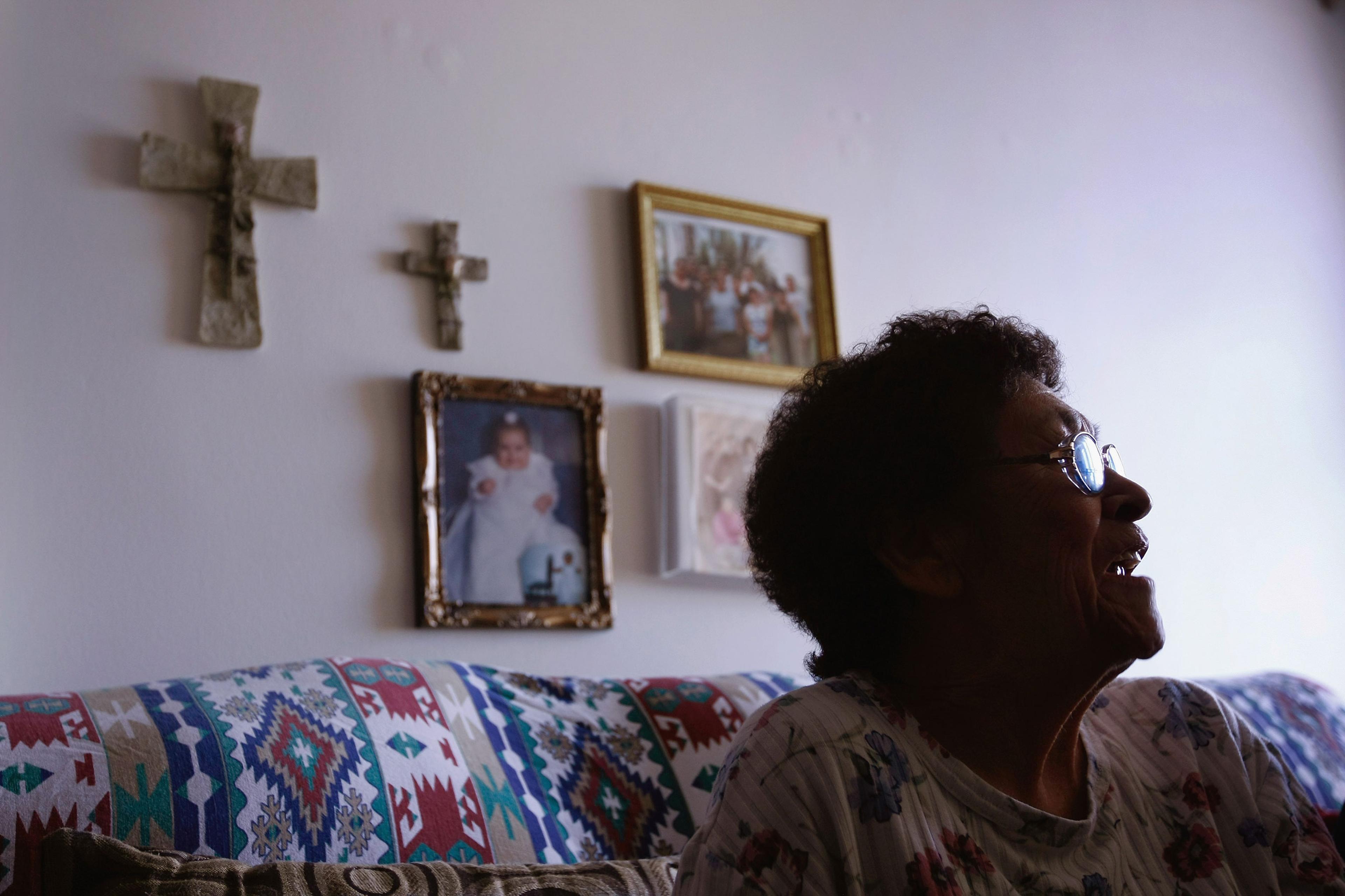 An elderly woman in profile smiling, seated on a patterned sofa with wall art and crosses in the background.