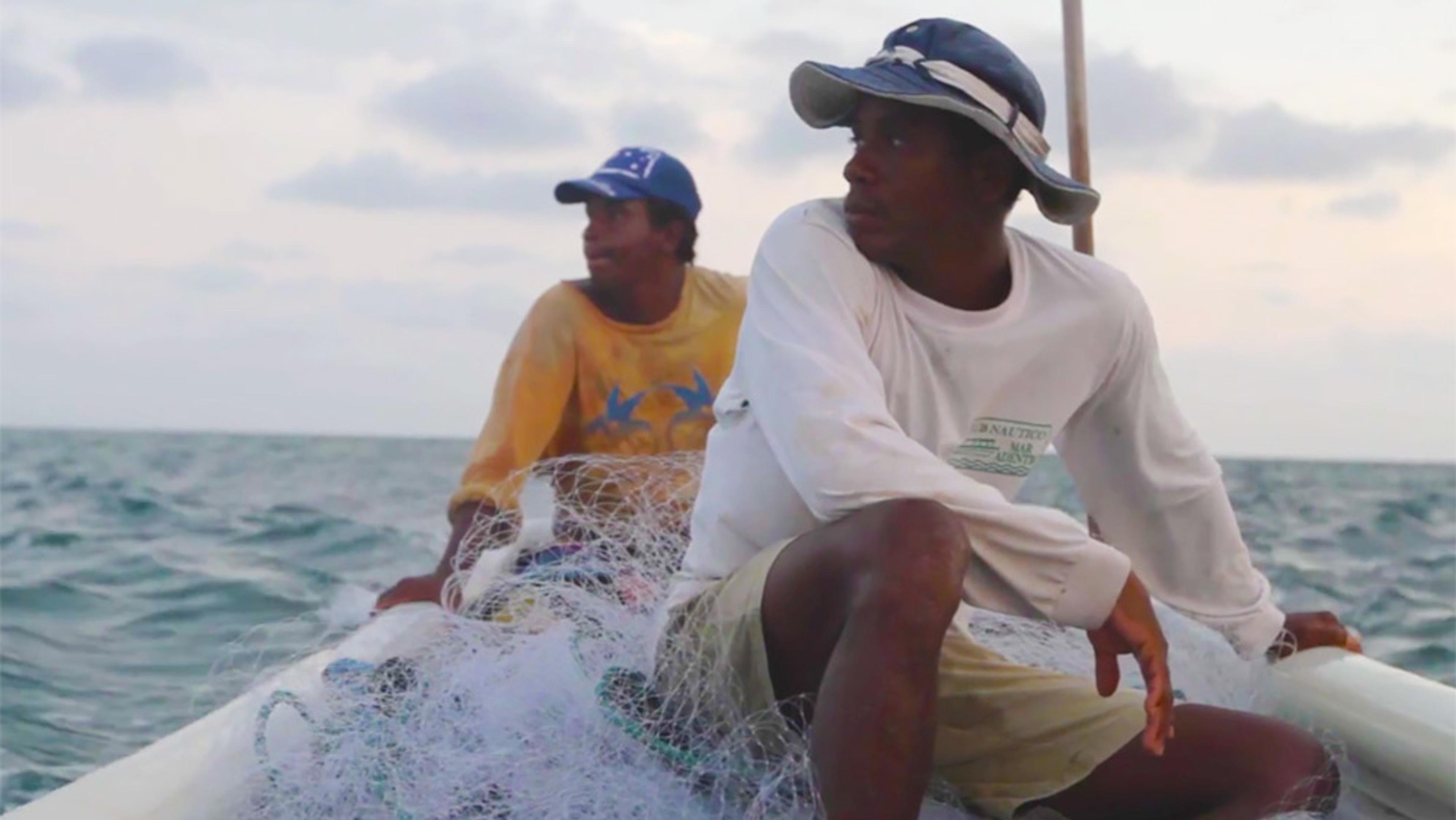 Two men on a boat at sea, wearing hats and casual clothes, surrounded by fishing nets, with a cloudy sky in the background.