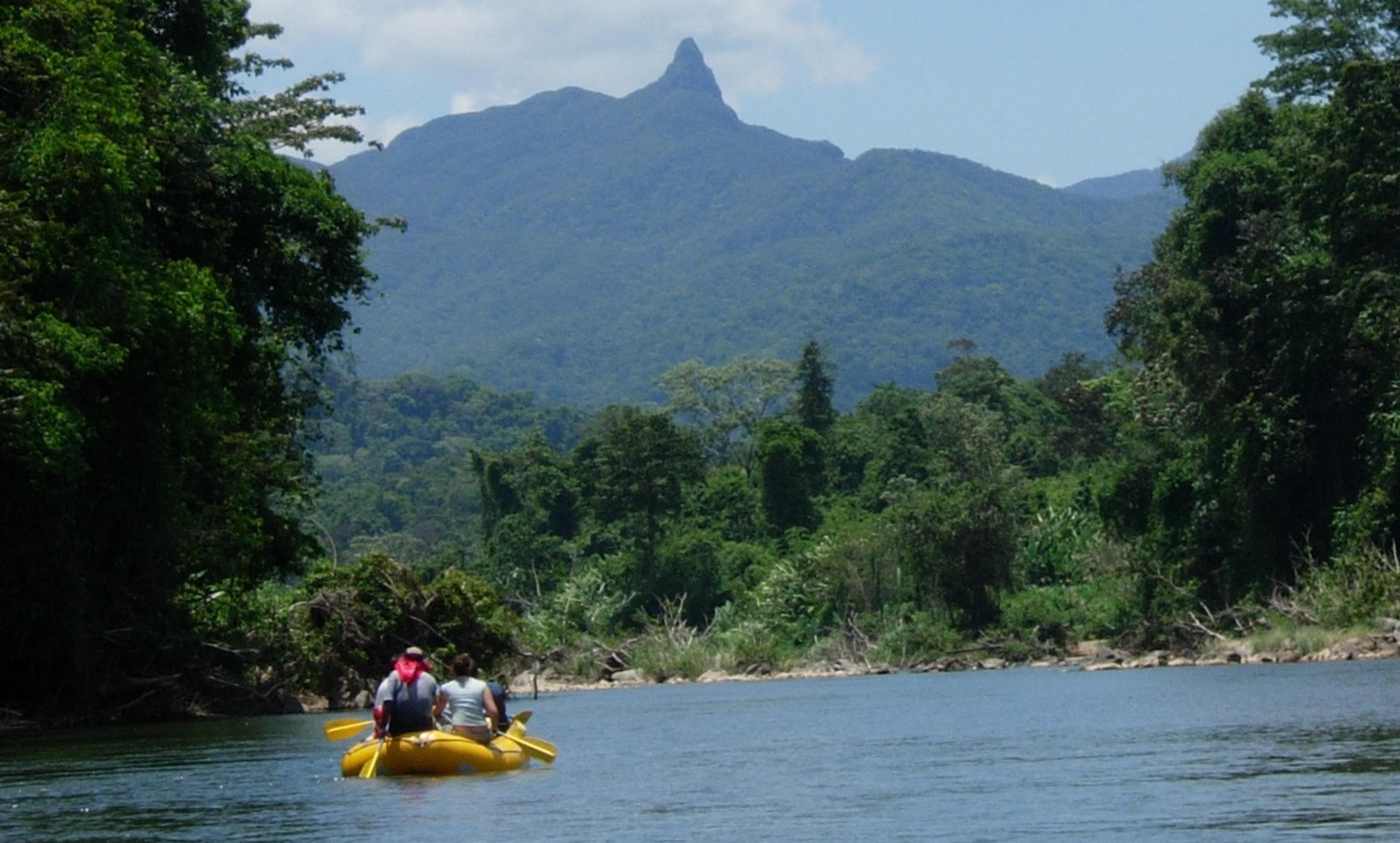 Three people in a yellow raft on a river surrounded by dense forest, with a mountain peak in the background.