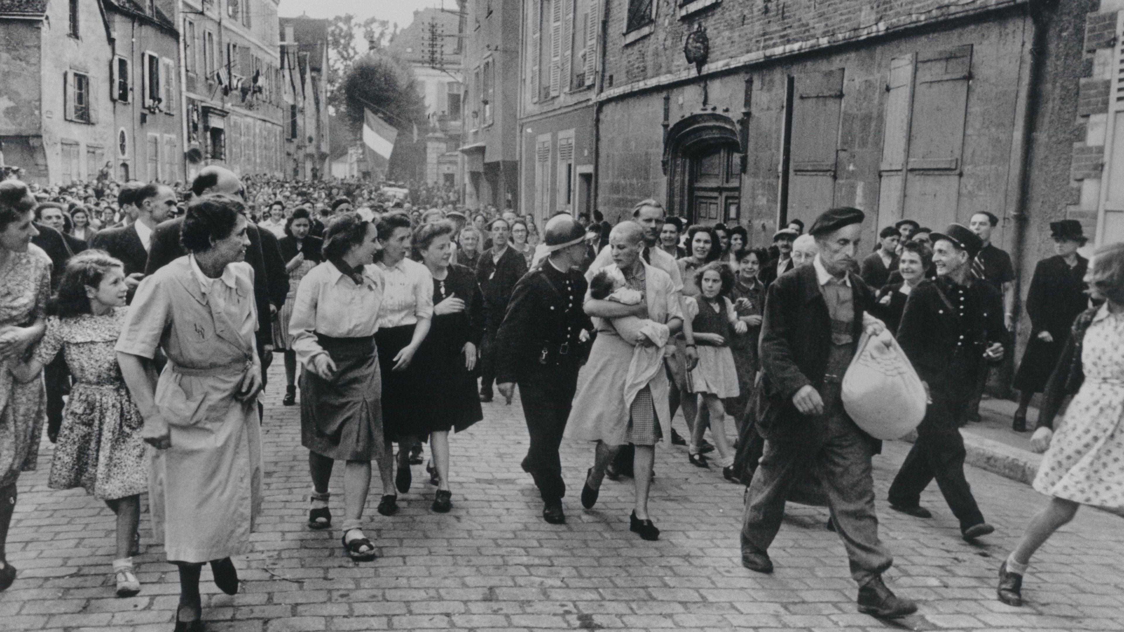Black and white photo of a crowd of people walking down a cobblestone street with buildings in the background.