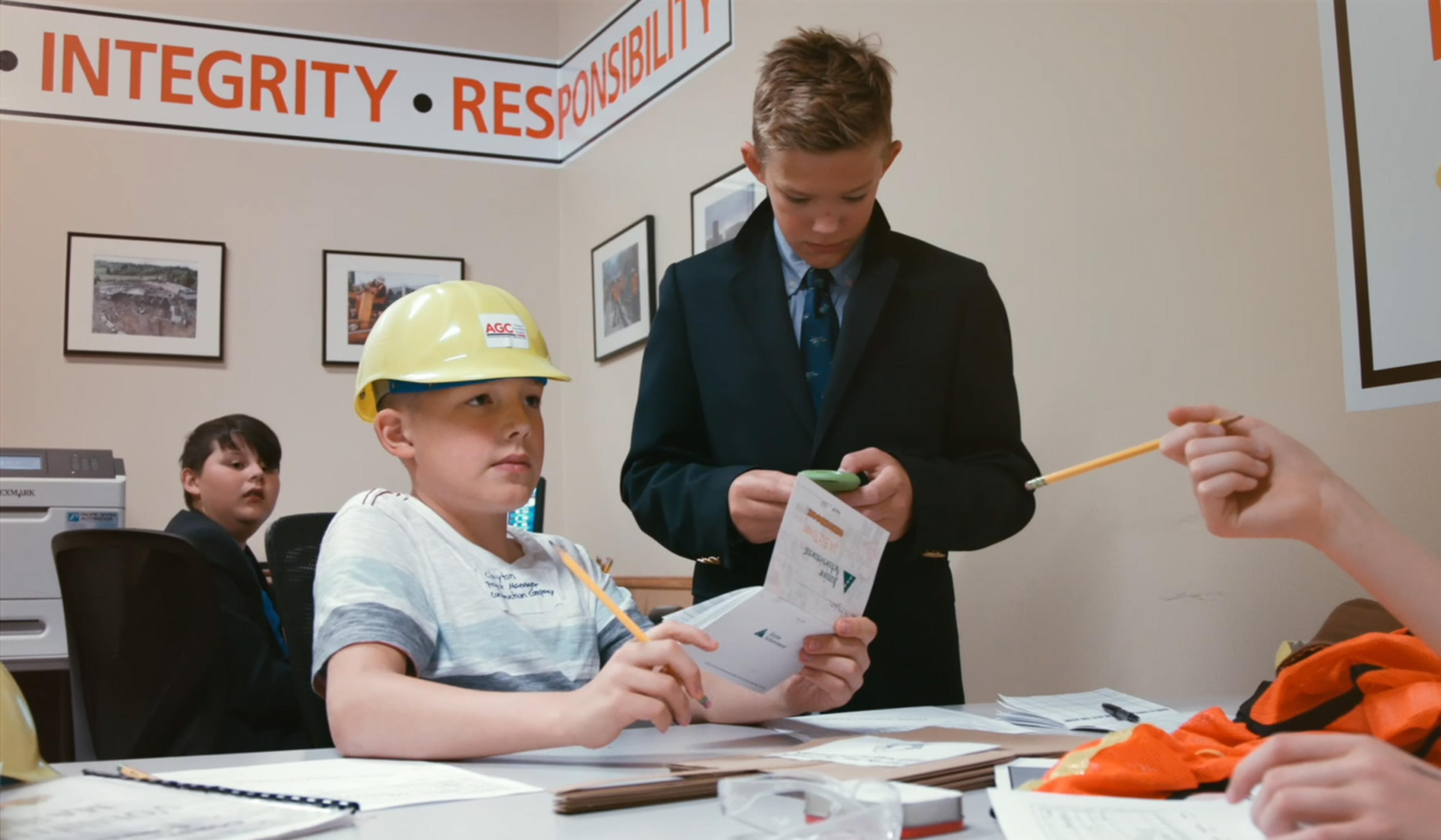 Children role-playing in an office; one wears a hard hat while another checks papers. Wall has “INTEGRITY” and “RESPONSIBILITY” signage.
