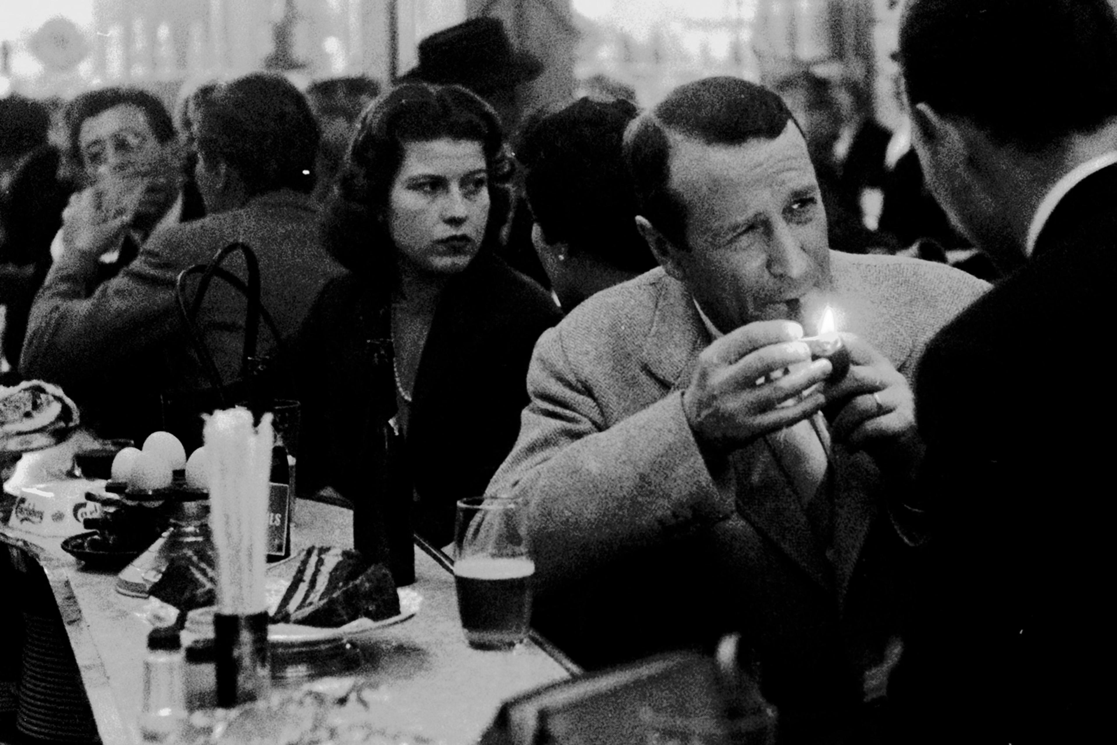 Black and white photo of people in a bustling bar, a man lighting a cigarette at a table with drinks and snacks.