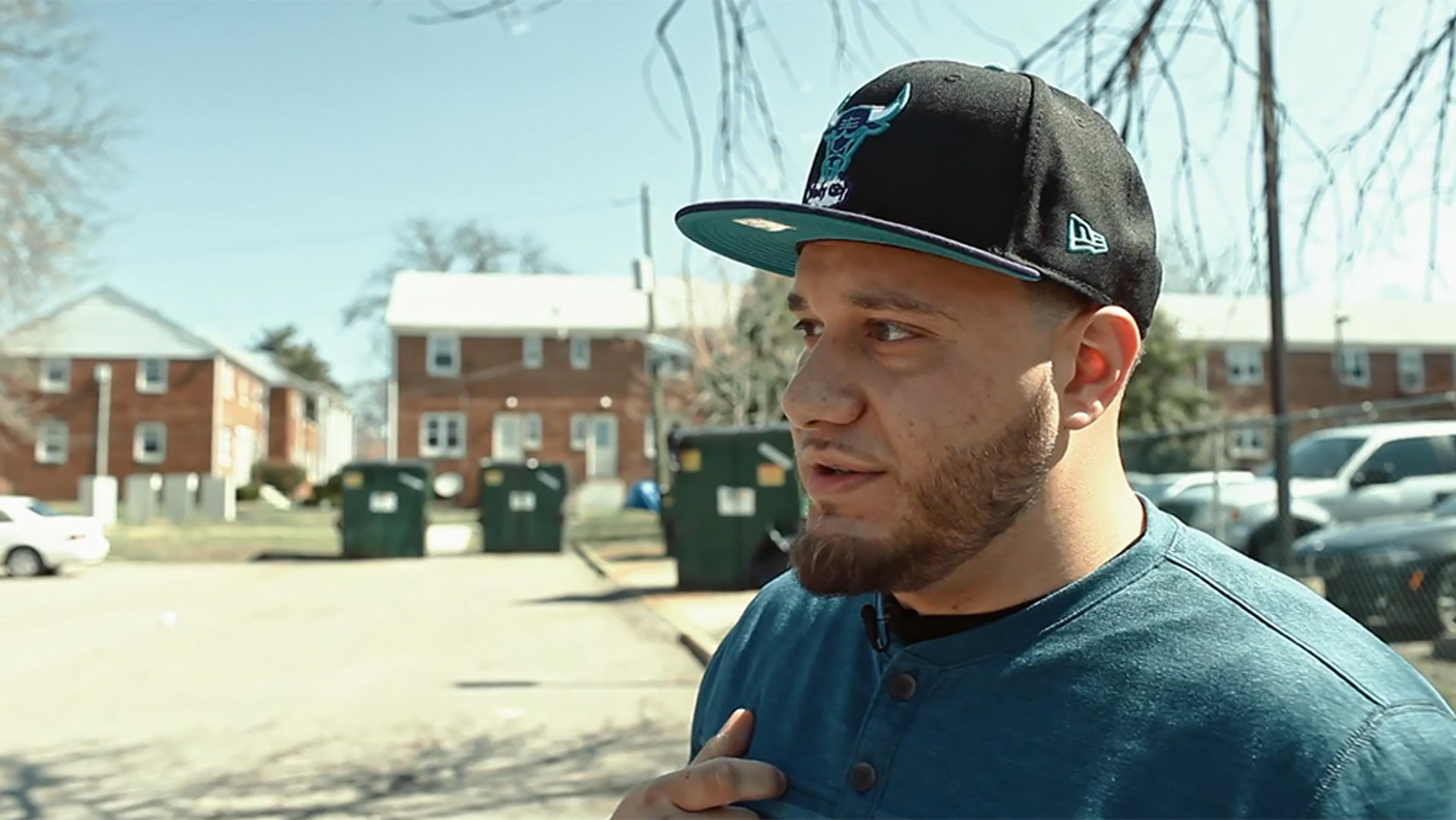 A man in a black cap in front of brick buildings lined with trees and several green bins on a sunny day.