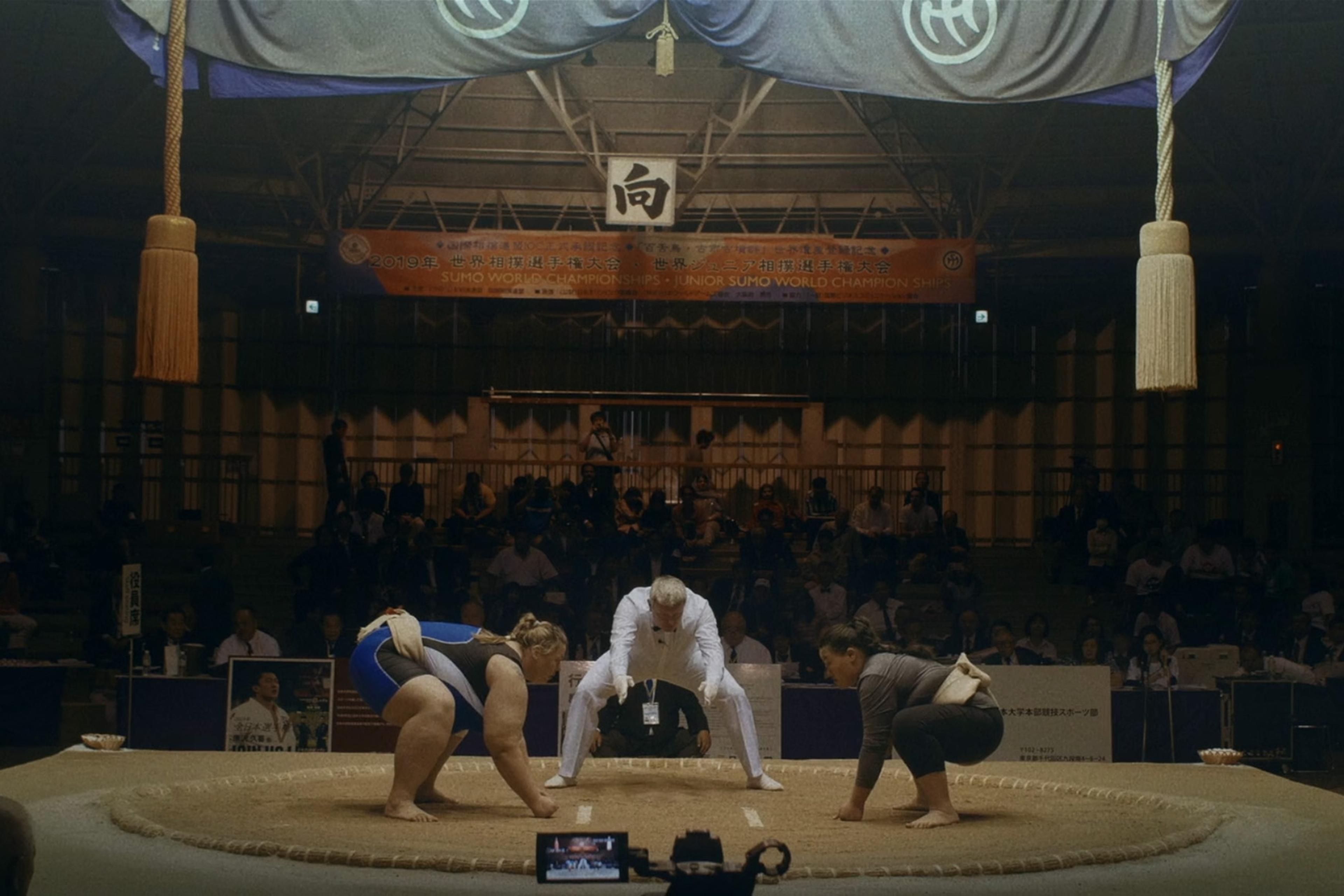 Two sumo wrestlers and a referee on a sumo ring during a match, with spectators in the background at the 2019 World Sumo Championships.