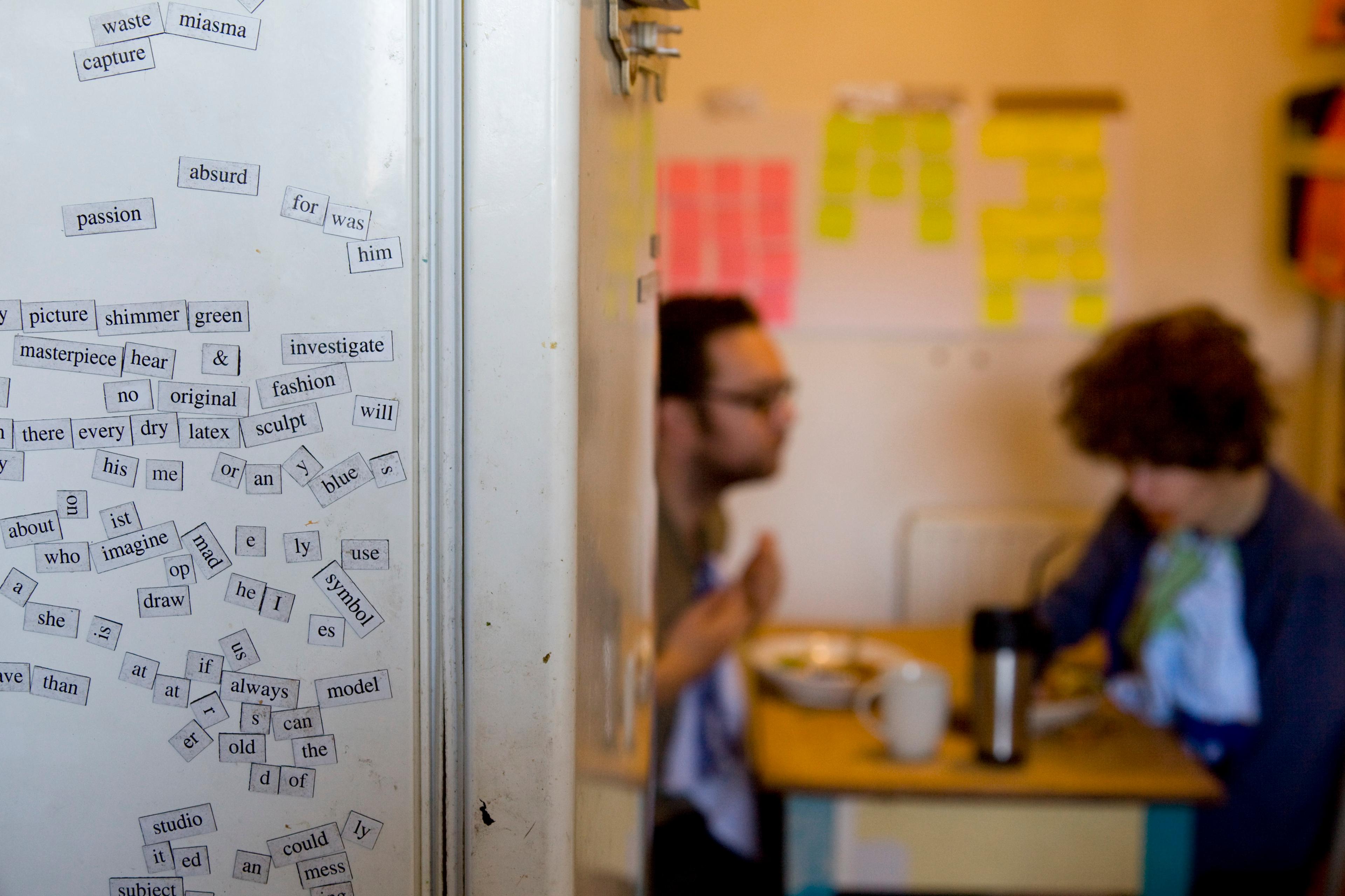 Photo of a kitchen with word magnets on a fridge and two people in the background at a table.