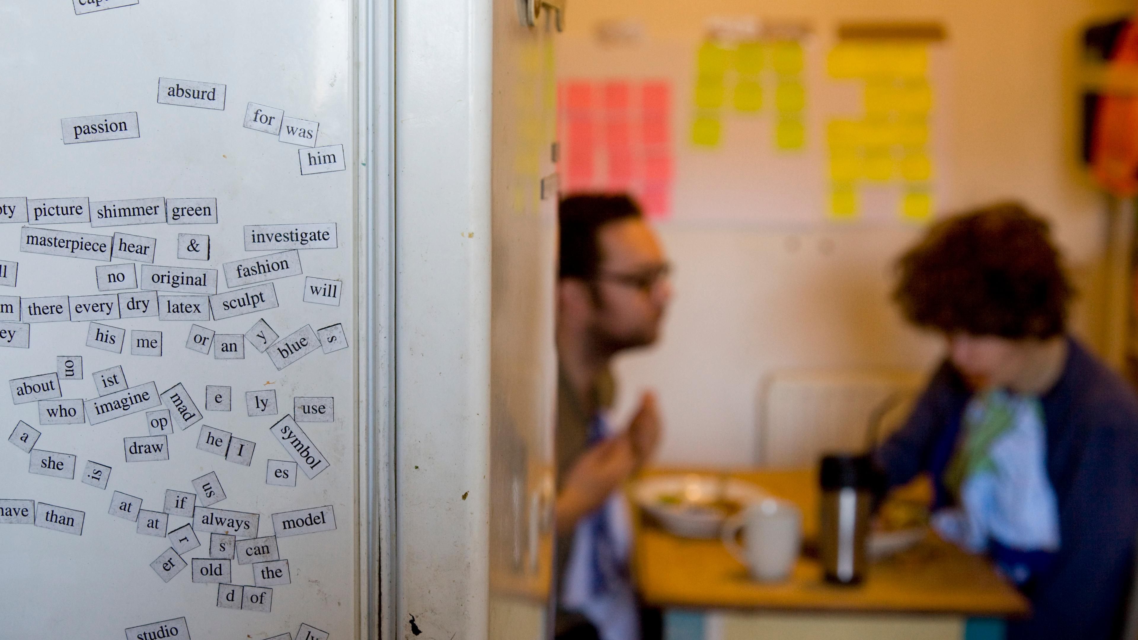 Photo of a kitchen with word magnets on a fridge and two people in the background at a table.