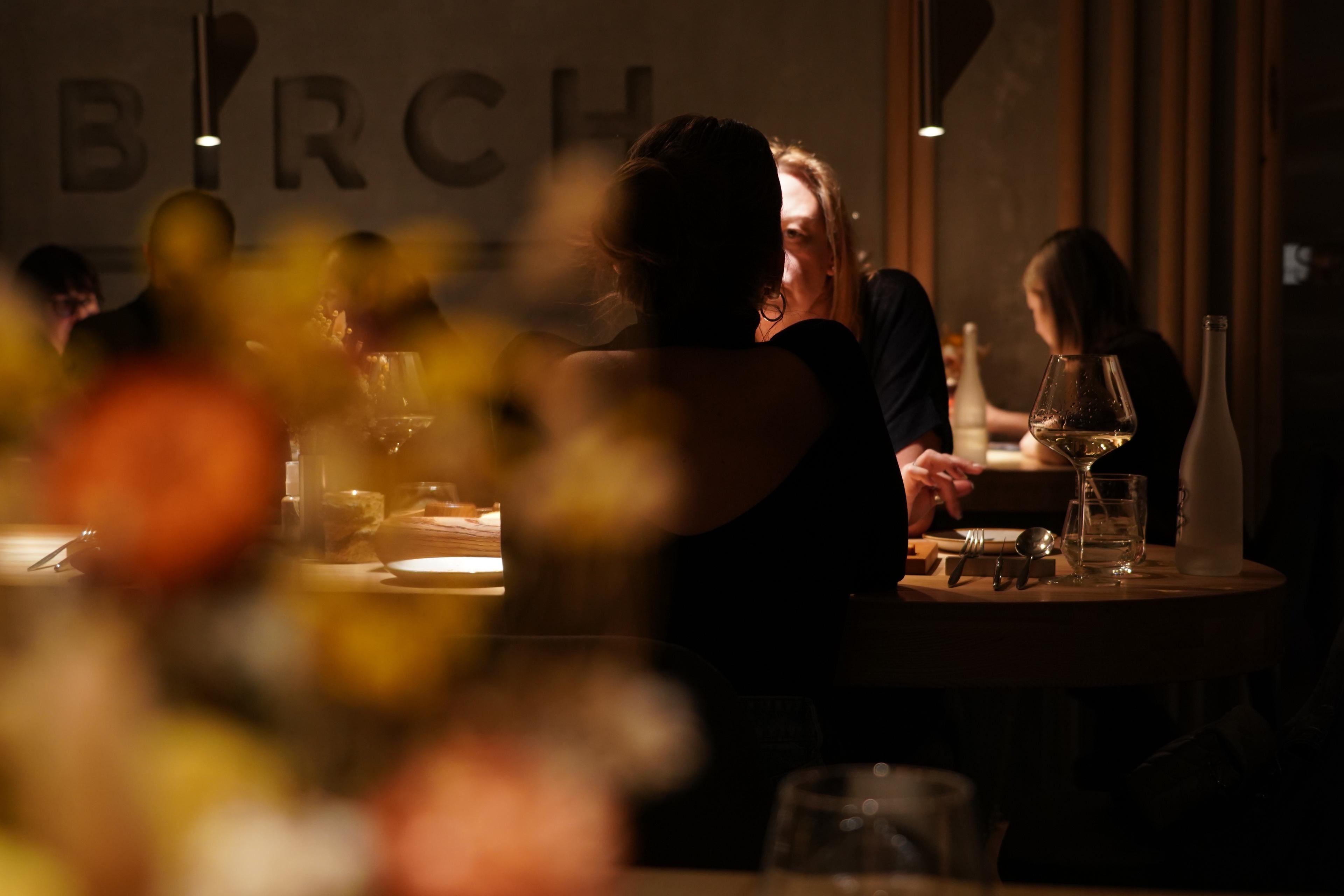 Photo of people dining in a dimly lit restaurant, blurred flowers in foreground, focus on background diners at a table.
