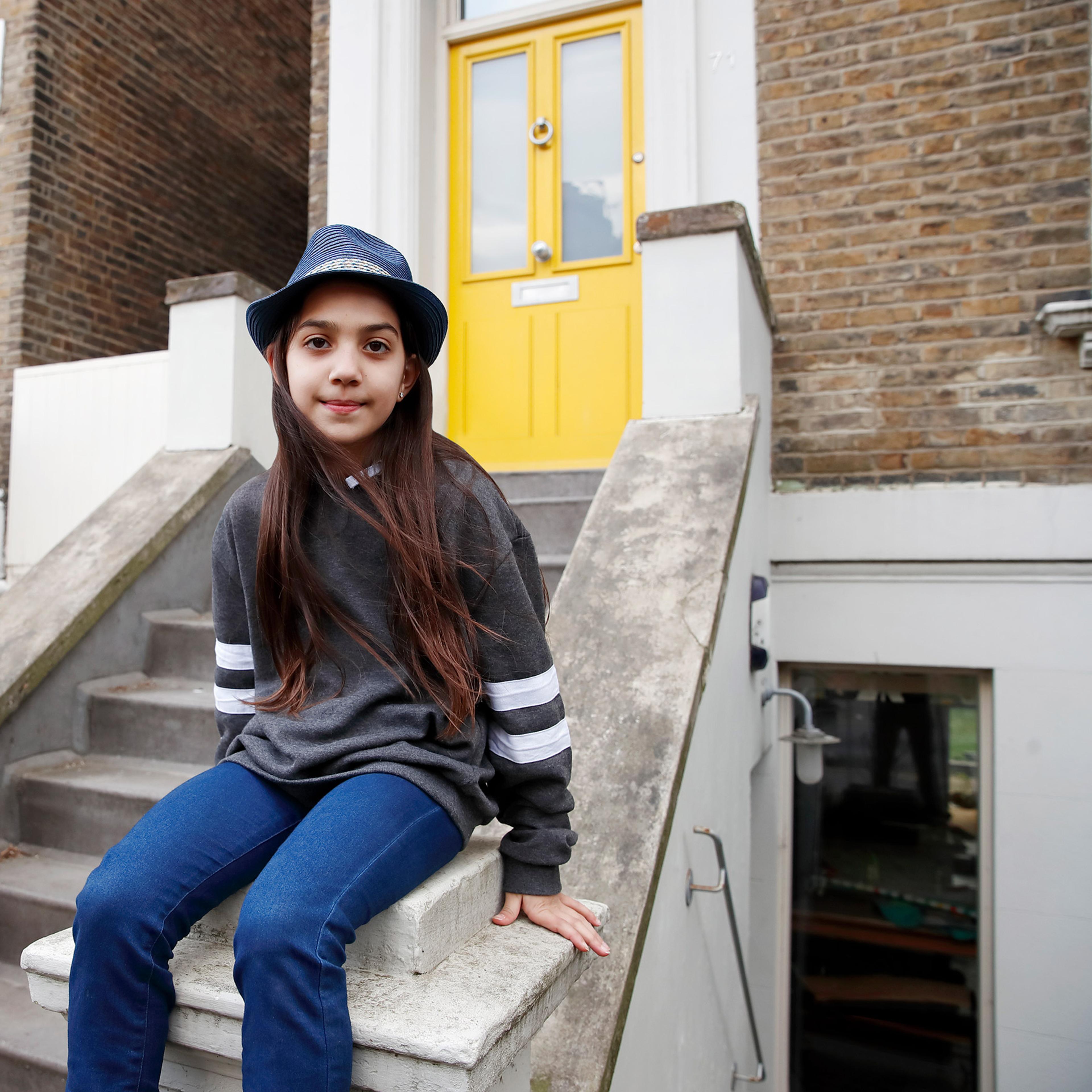 A young teenage girl wearing a trilby hat sits on the steps leading up to a yellow front door
