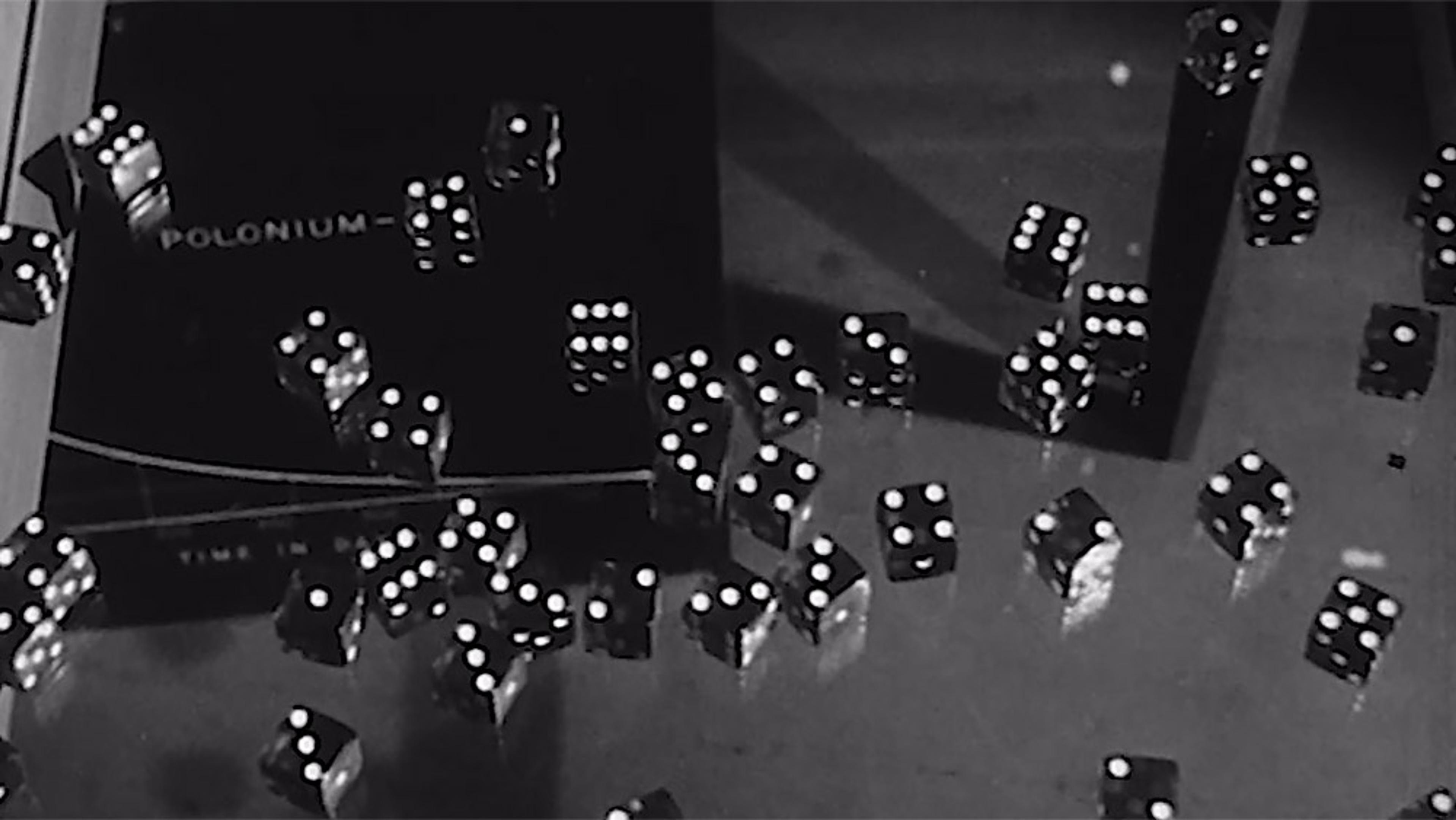 Black and white photo of multiple six-sided dice scattered on a surface. Some dice are mid-air. A book with “Polonium” is partially visible.