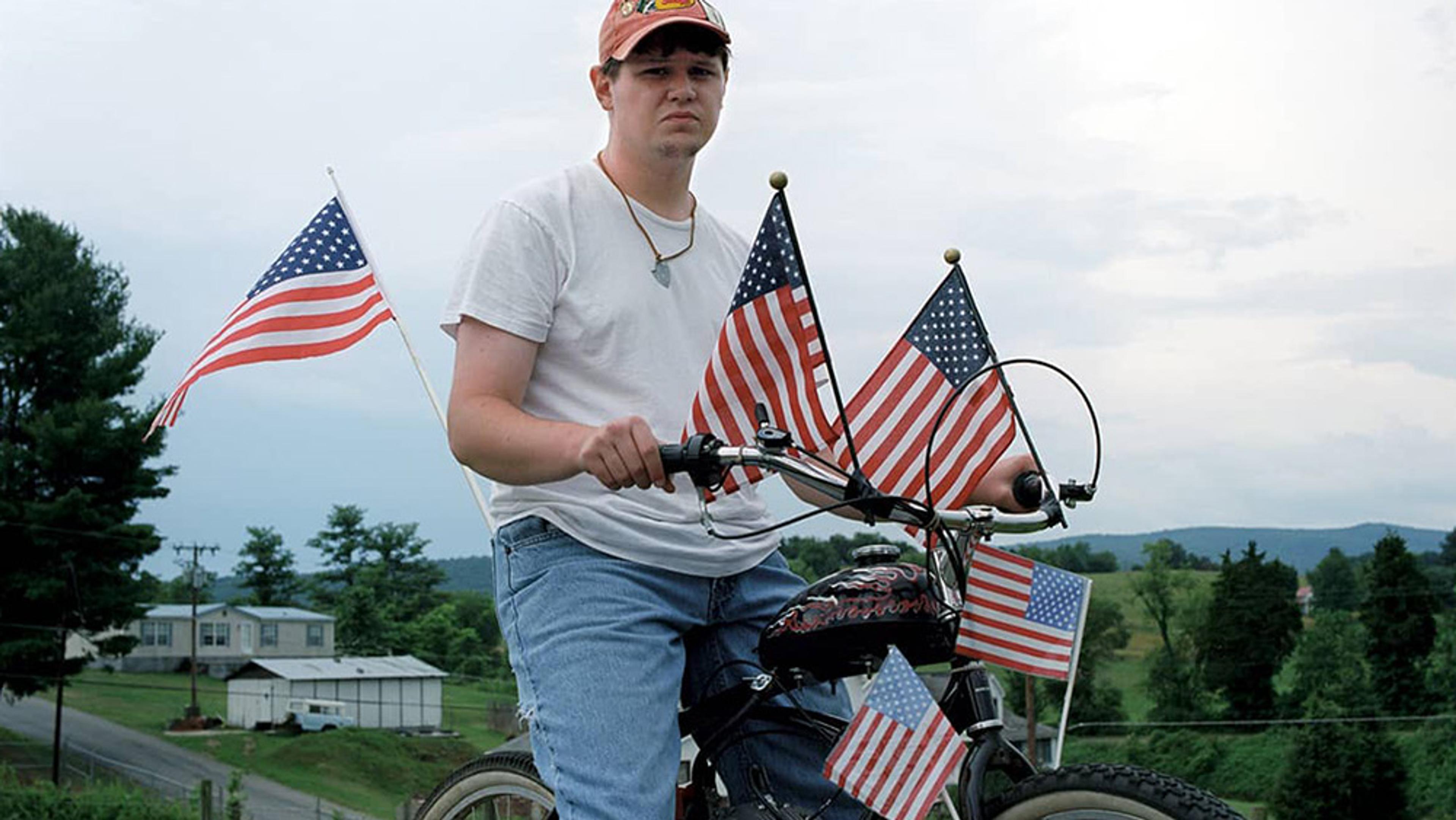 A man on a decorated bike with US flags in a rural area wearing a cap, white shirt and jeans with a cloudy sky background