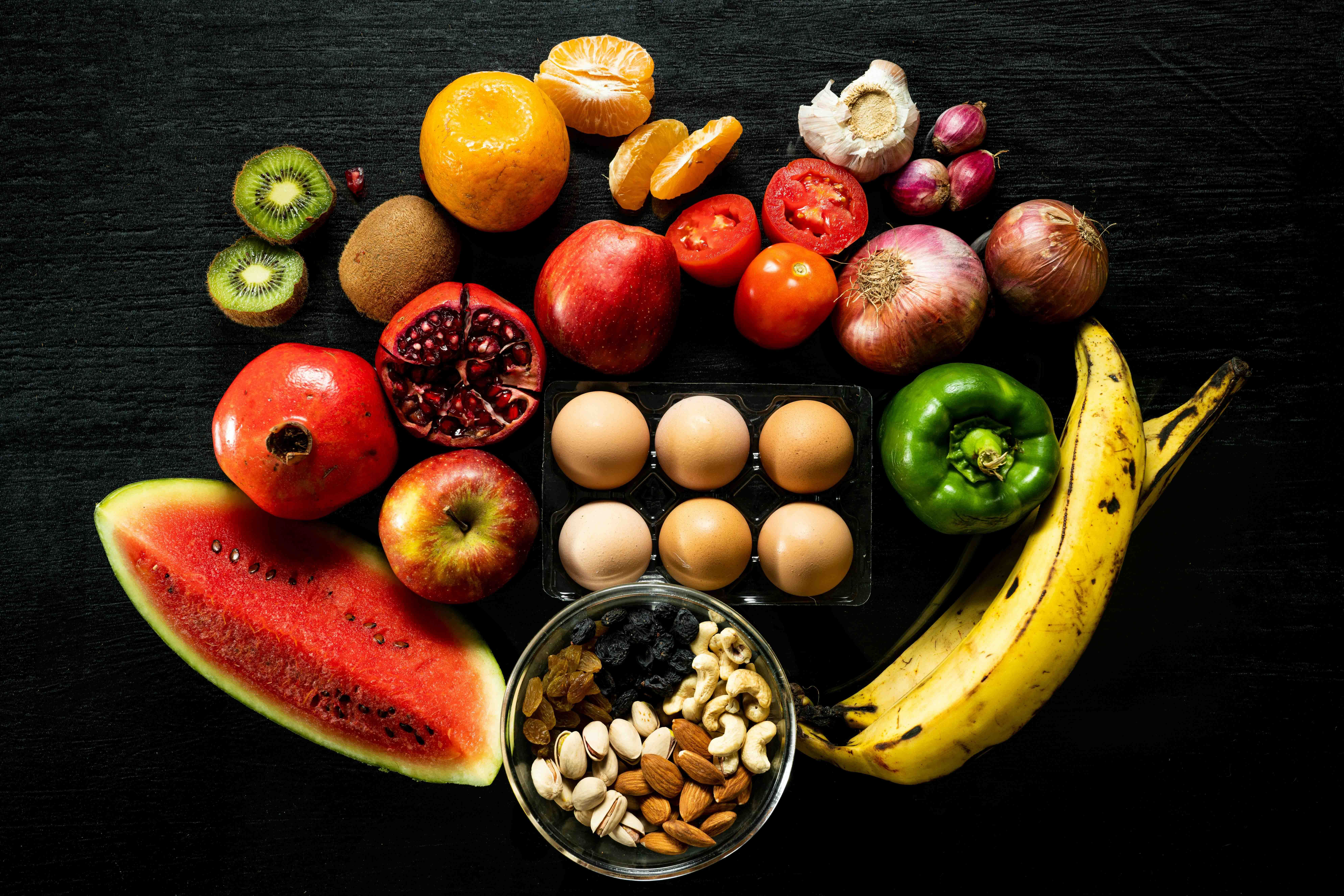 Photo of assorted fruits, vegetables, a tray of eggs and a bowl of nuts on a black background.