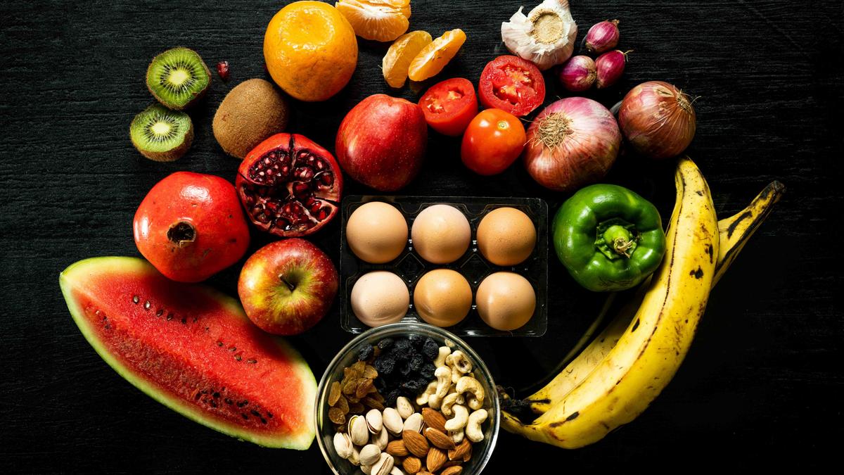 Photo of assorted fruits, vegetables, a tray of eggs and a bowl of nuts on a black background.
