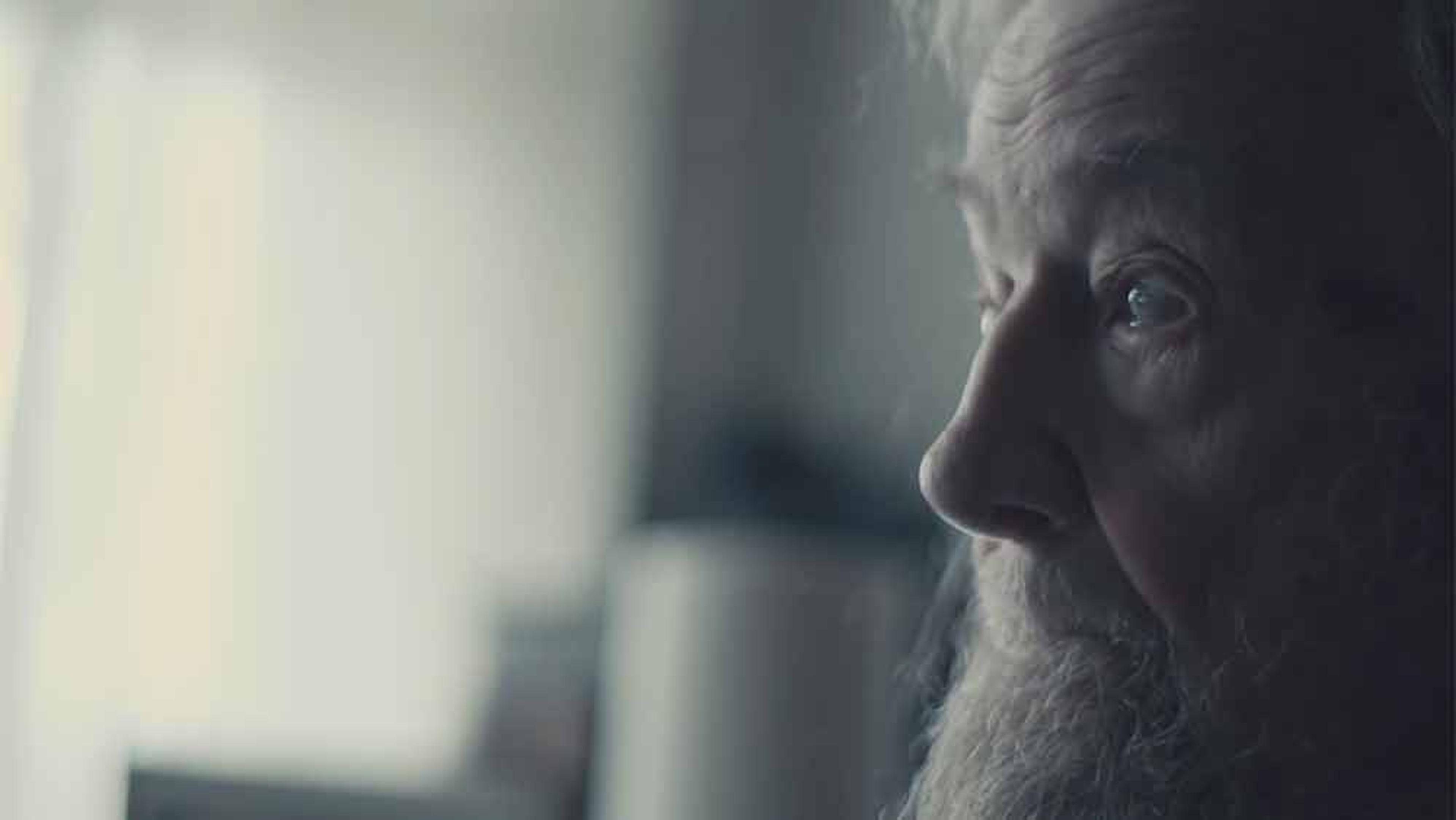 An elderly man with a beard looking to the right, in soft lighting with a blurred background.