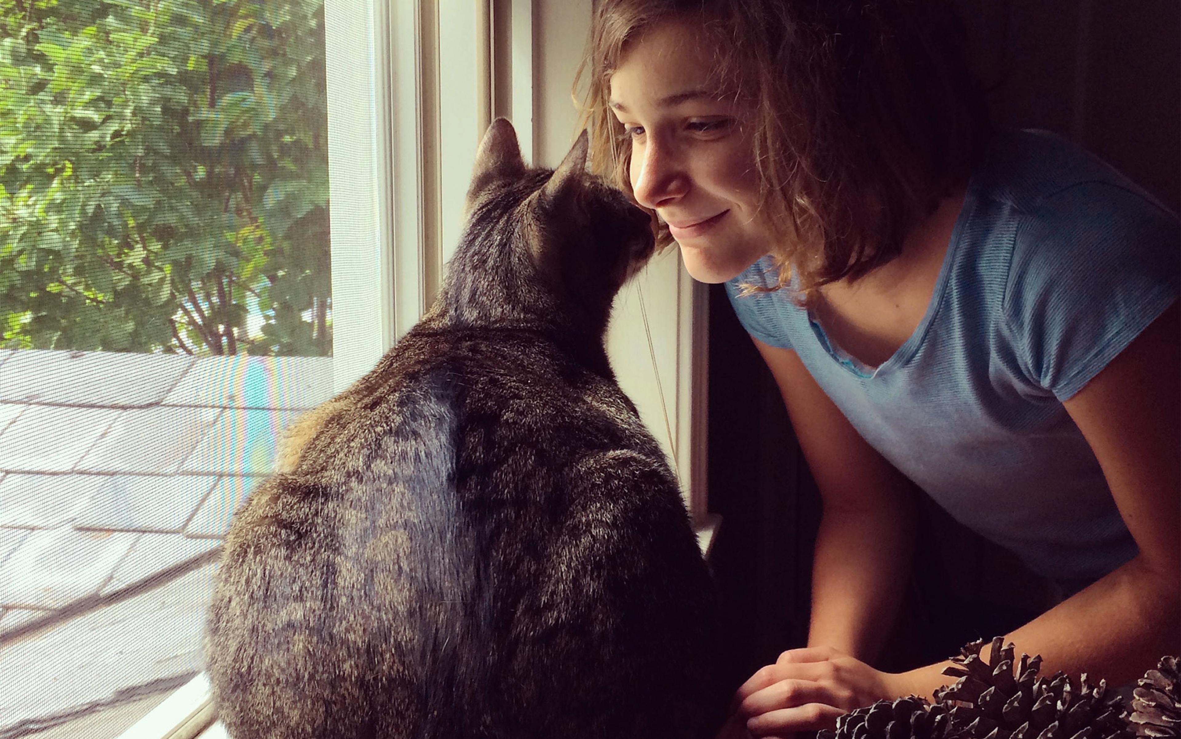 A smiling young girl and a cat, nose to nose, by a window sill, with pinecones beside them