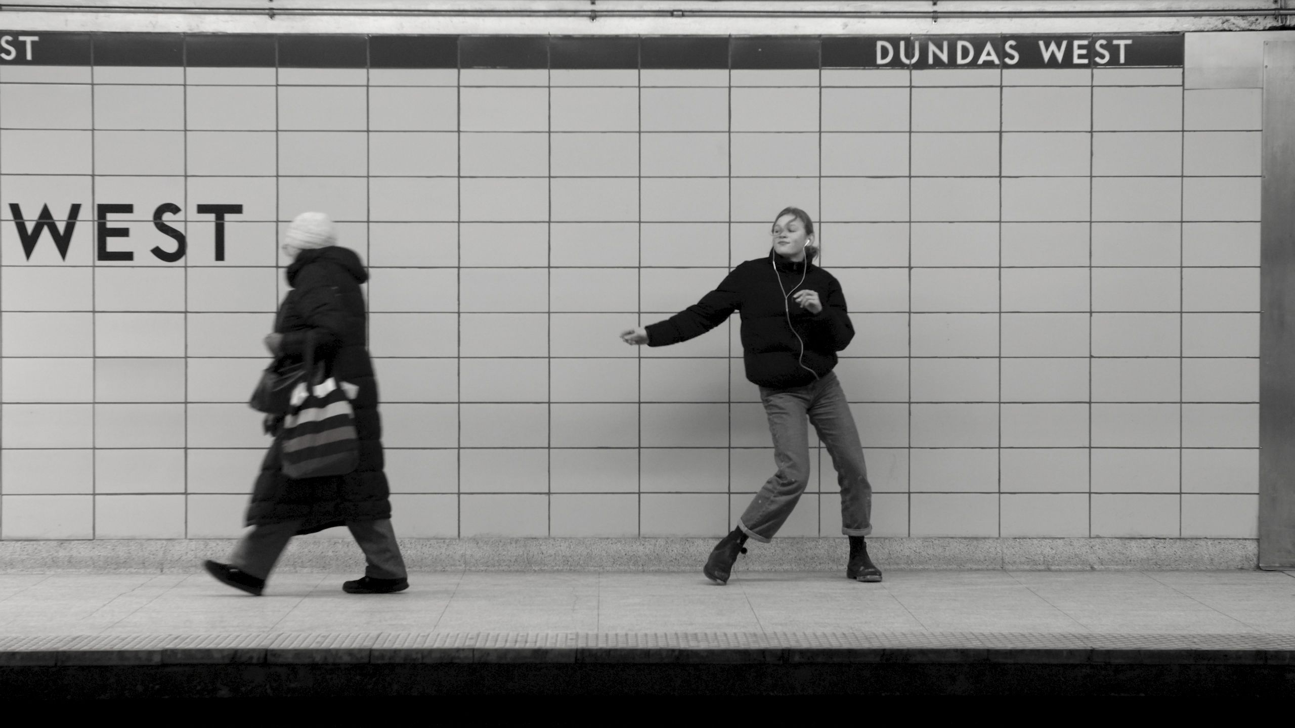 Black and white photo of two people in a subway station, one walking left, another dancing, station name Dundas West.