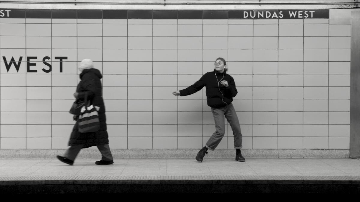 Black and white photo of two people in a subway station, one walking left, another dancing, station name Dundas West.