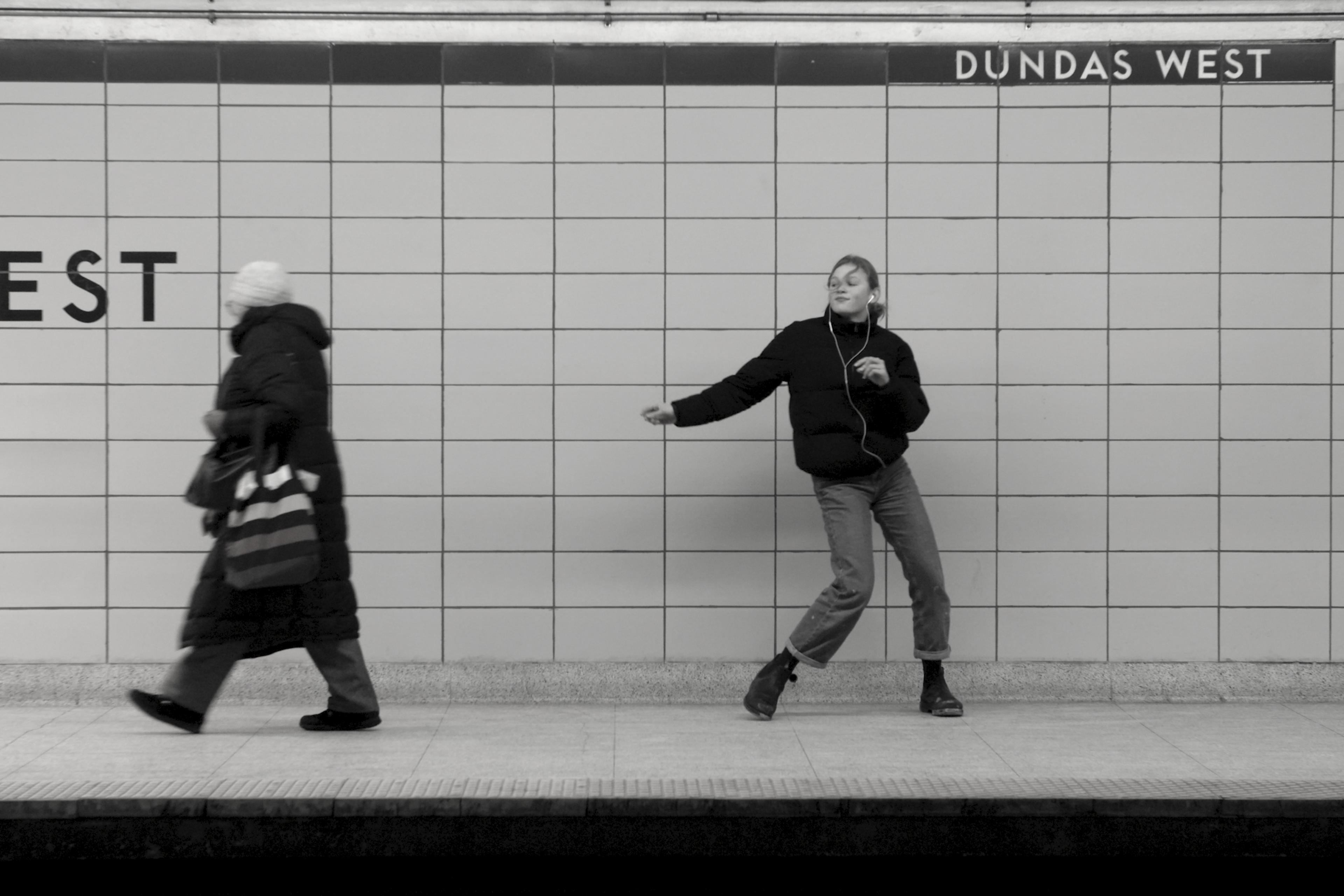 Black and white photo of two people in a subway station, one walking left, another dancing, station name Dundas West.