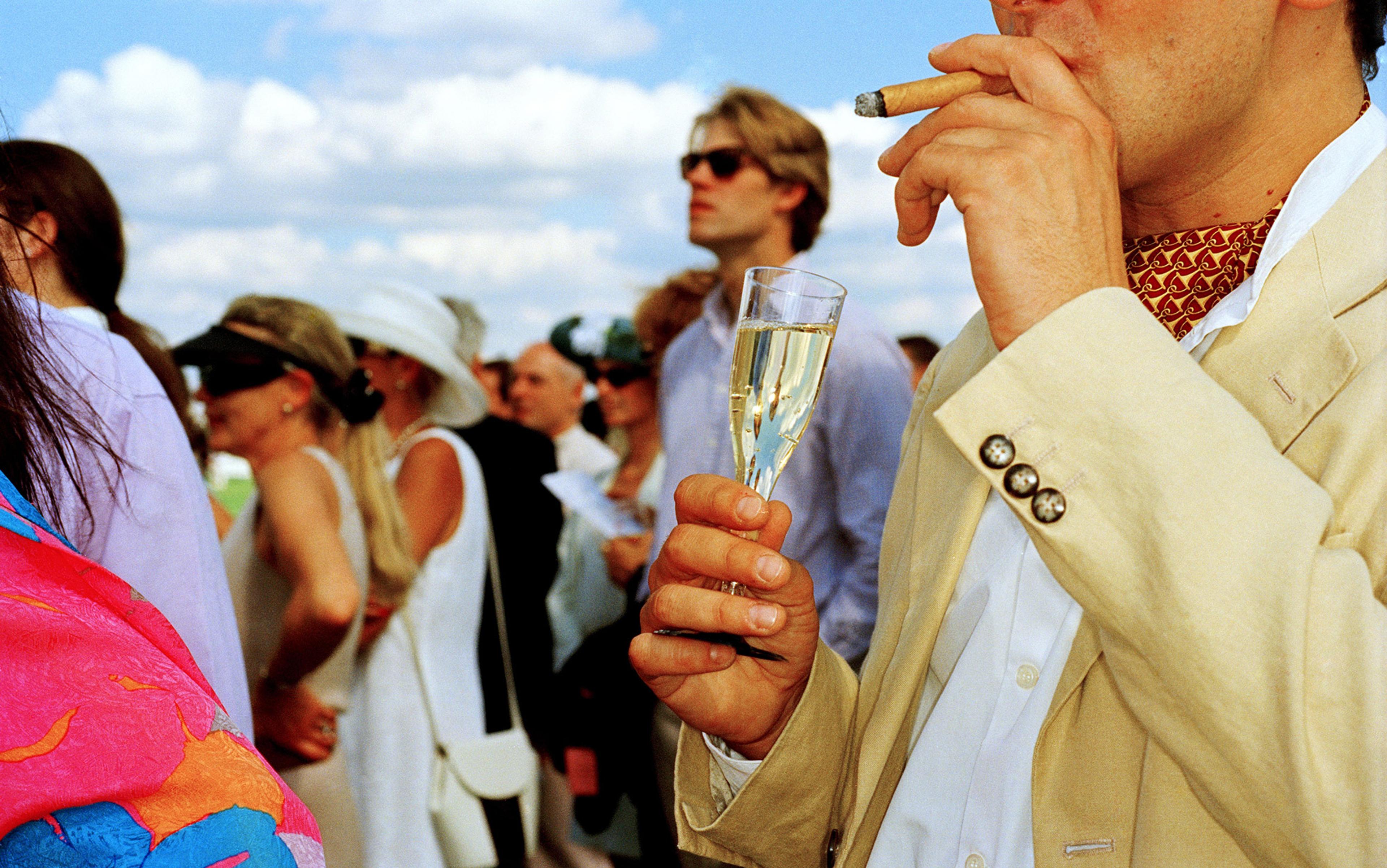 A man smoking a cigar and holding a champagne flute at an outdoor event, with people in the background on a sunny day.