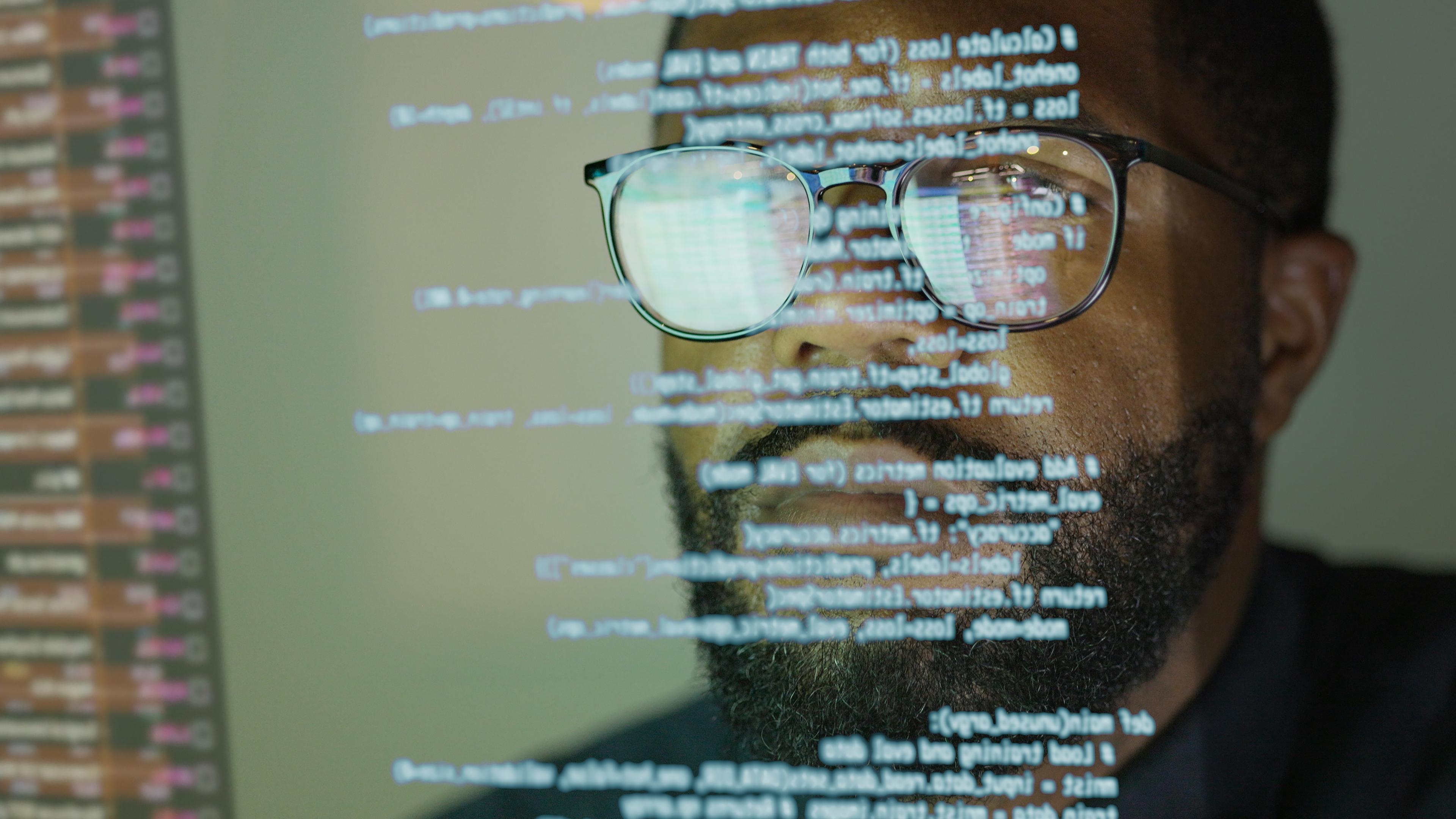 Photo of a person with code reflecting on glasses, focused on a computer screen displaying programming text.