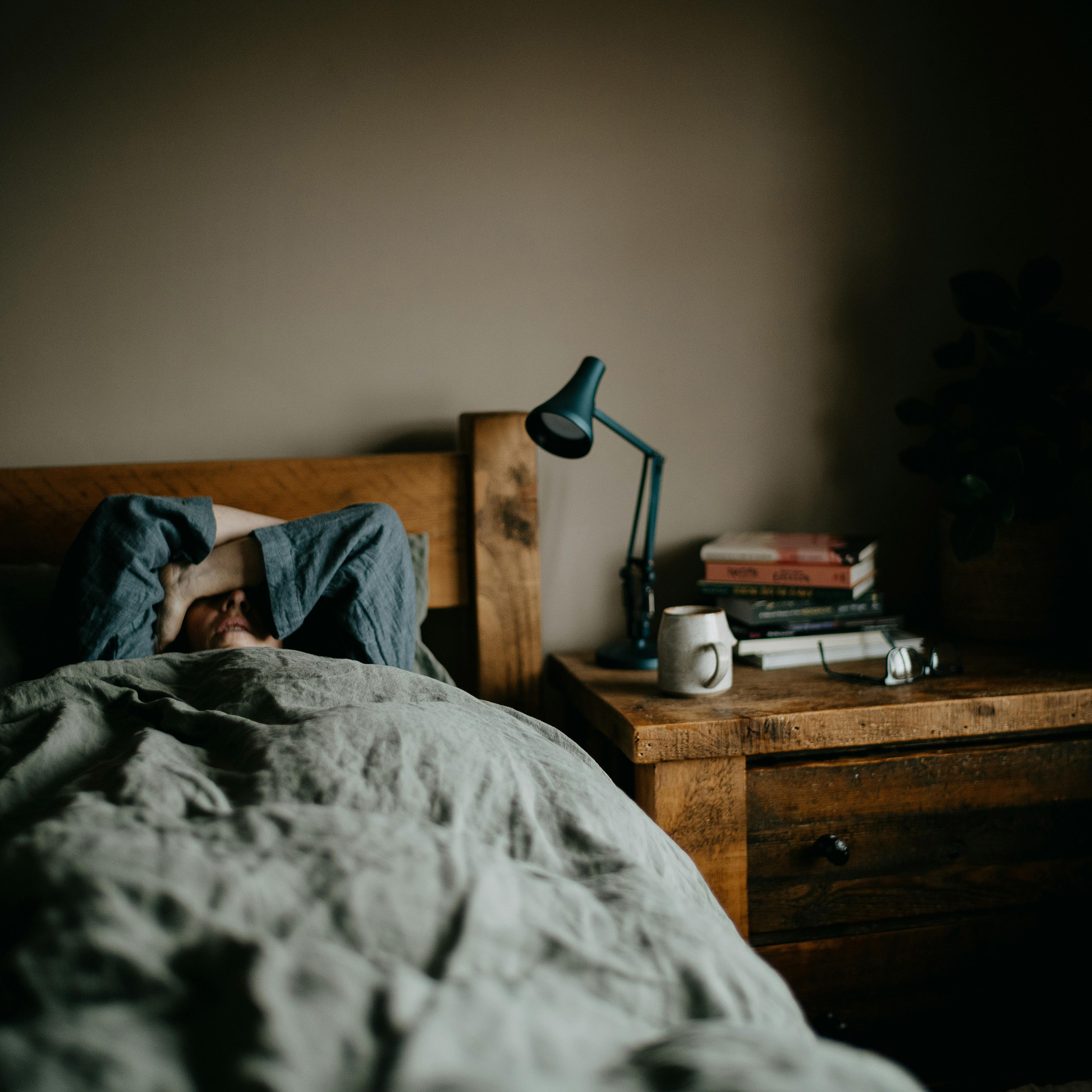 A person lying in bed under a duvet, arms covering face; bedside table with books, a lamp, a mug and reading glasses.