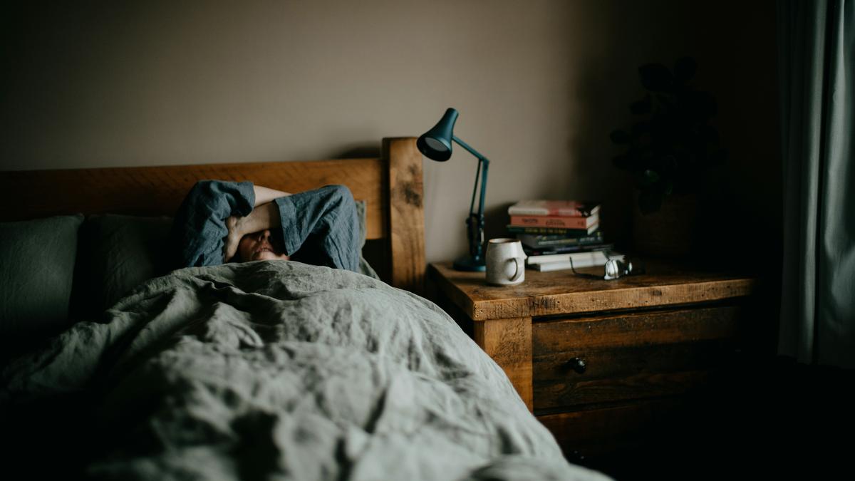 A person lying in bed under a duvet, arms covering face; bedside table with books, a lamp, a mug and reading glasses.