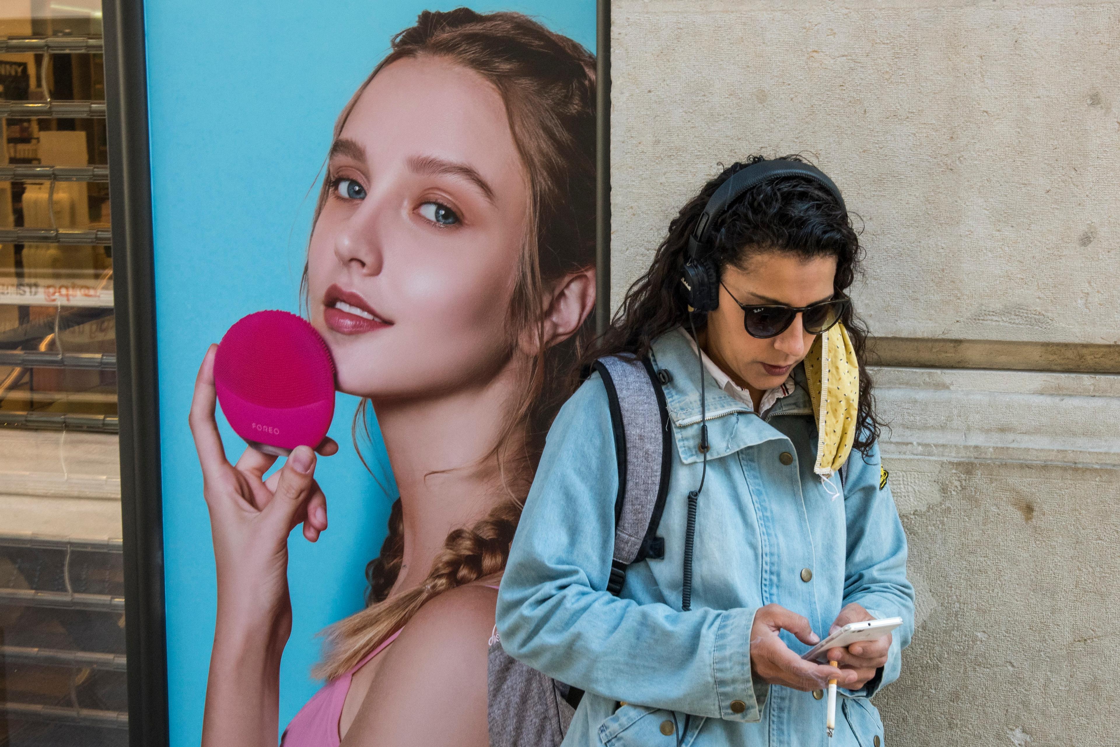 Photo of a woman with headphones looking at her phone next to an advert showing a model holding a pink facial brush.